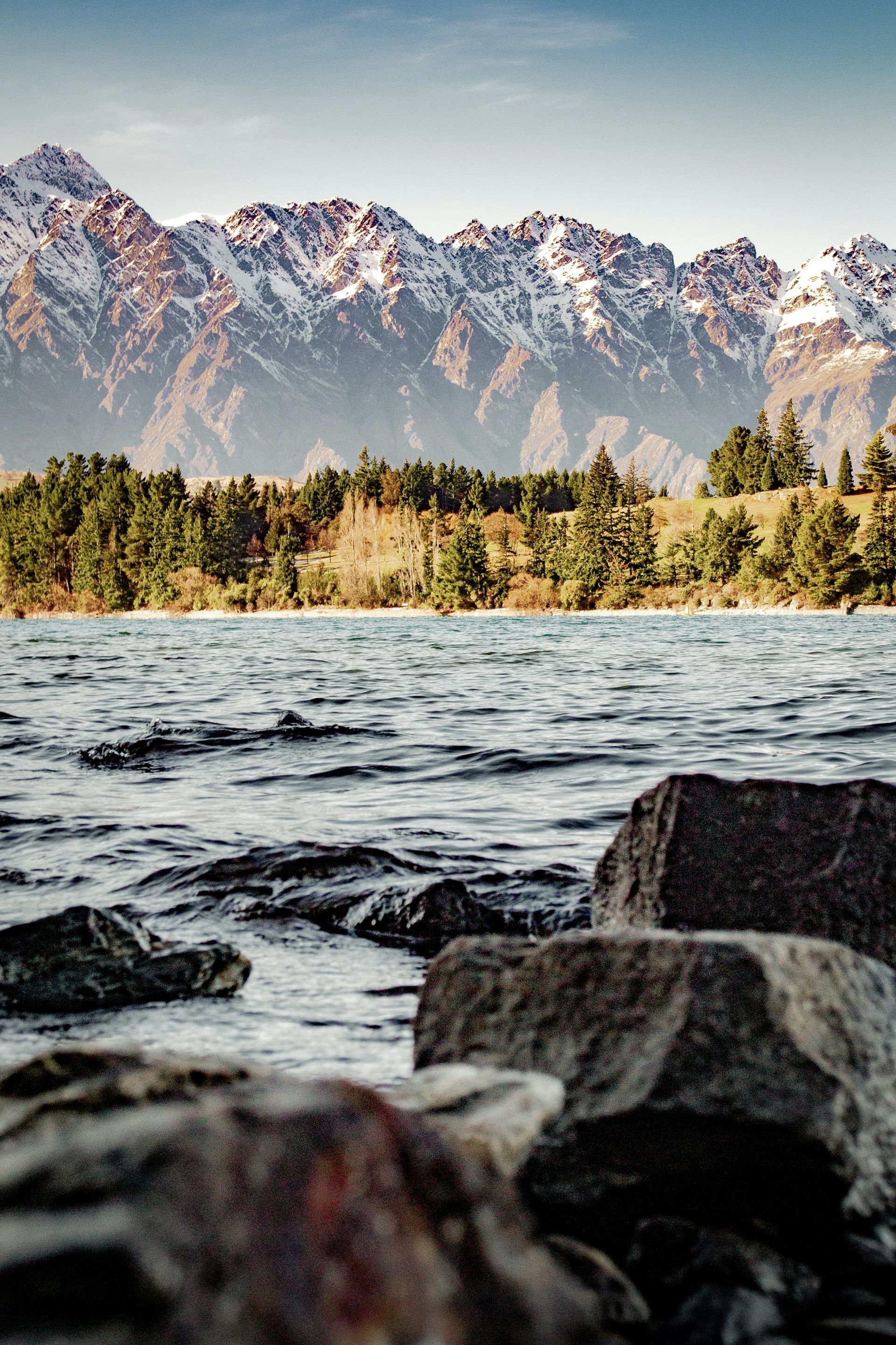 A lake with mountains in the background and rocks in the foreground in Queenstown, New Zealand.