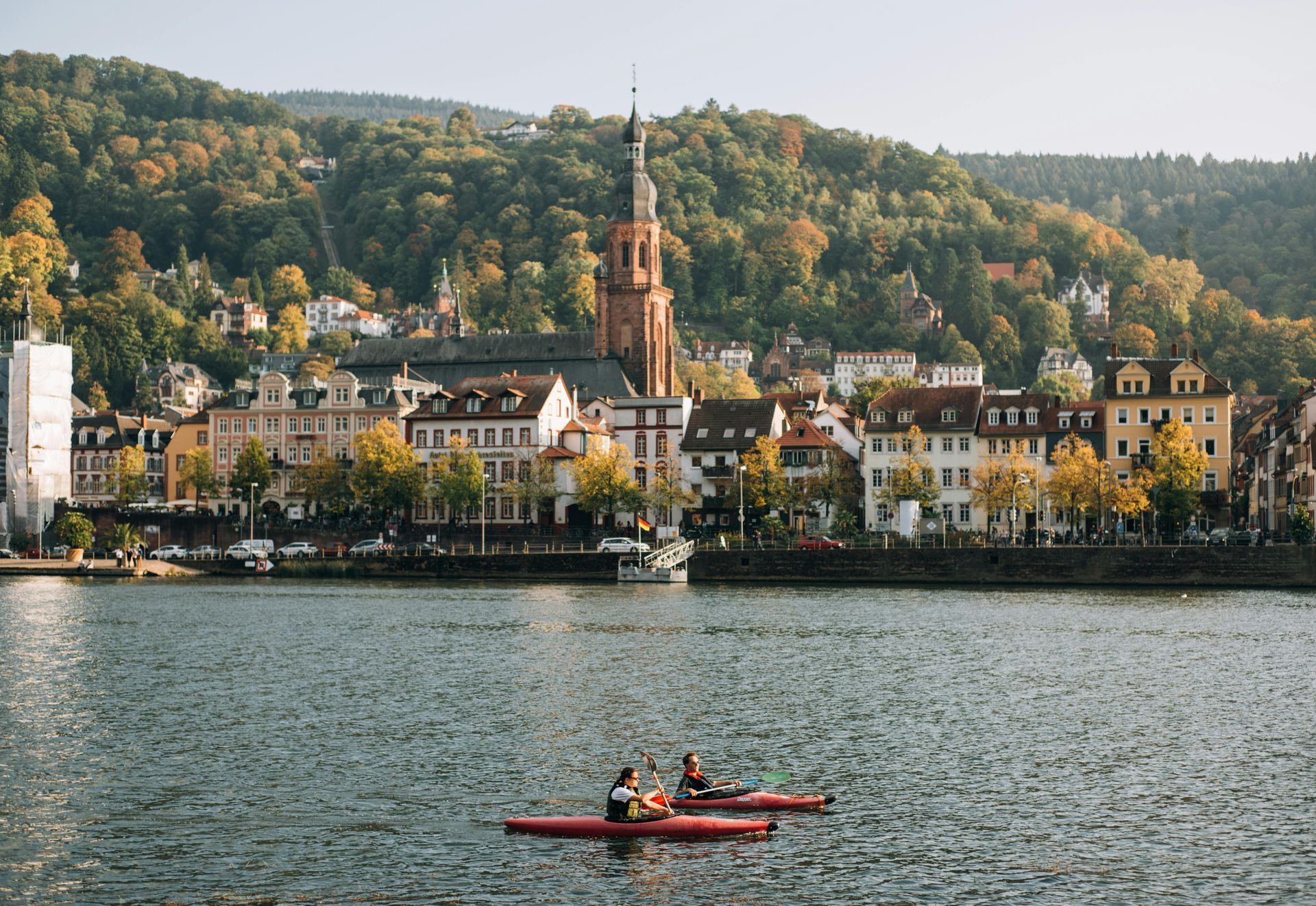 Two people are paddling kayaks on a lake with Heidelberg, Germany in the background.