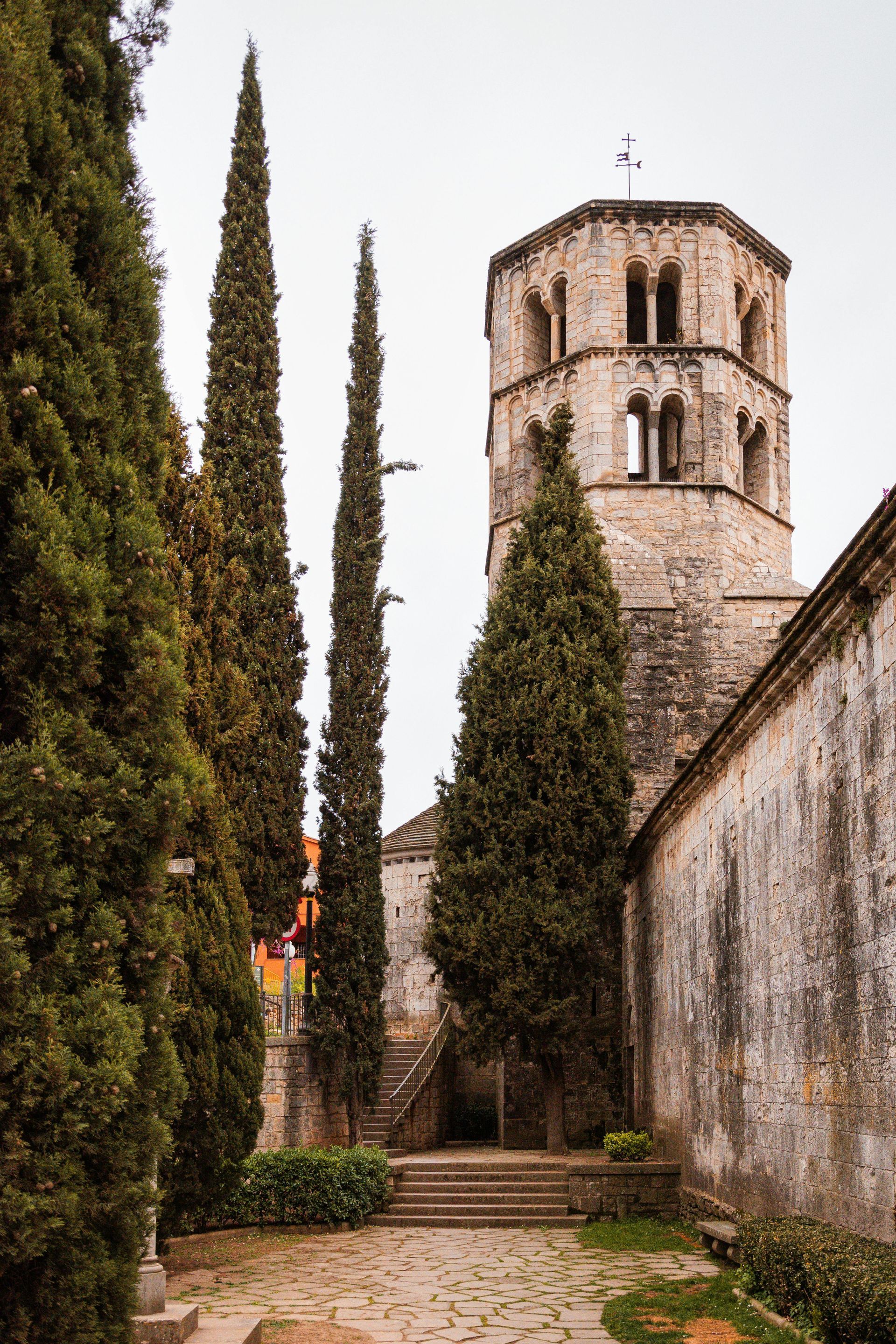 A tower with a cross on top of it is surrounded by trees and a stone wall in Girona, Spain