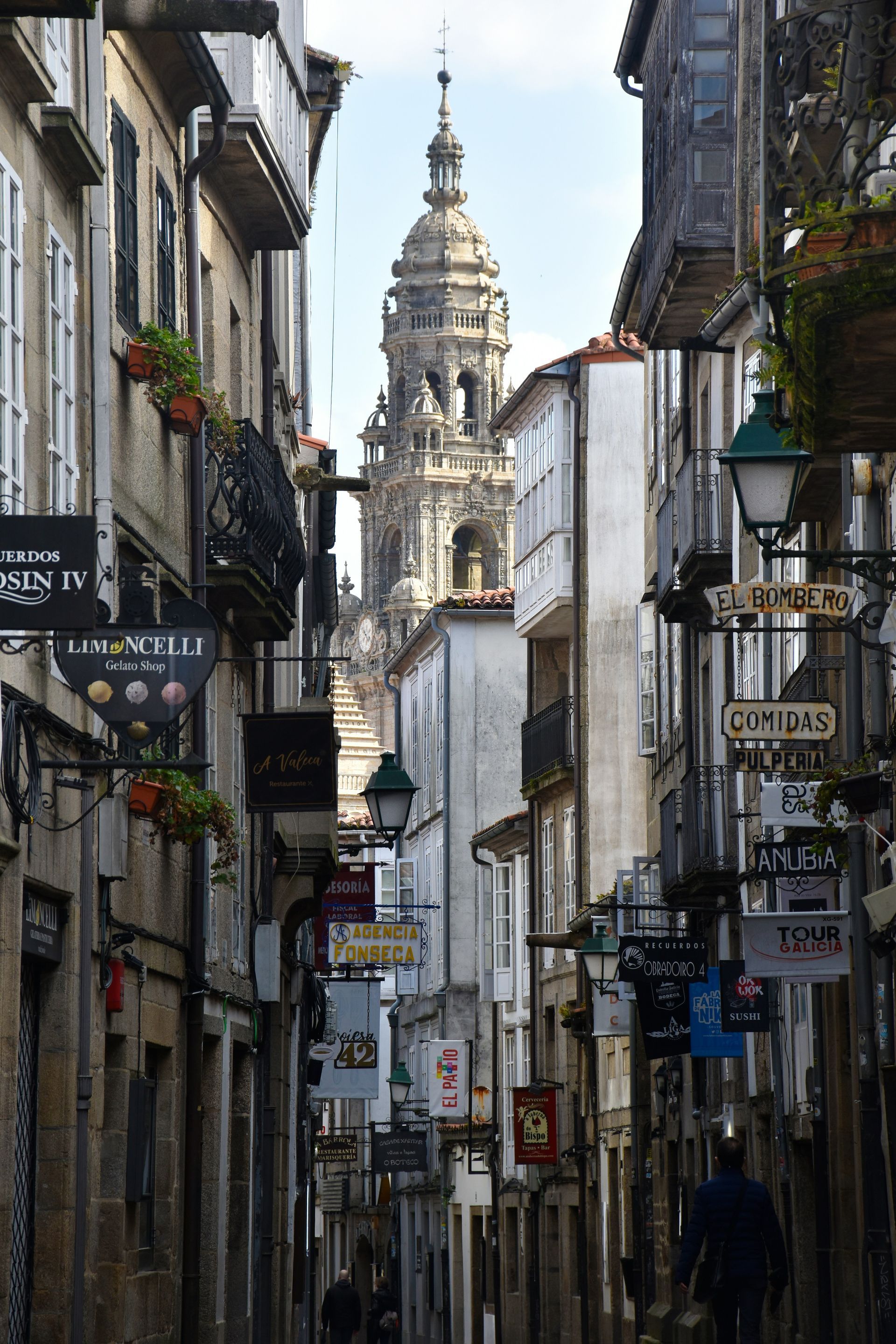 Narrow street in Santiago de Compostela with a tall cathedral tower at the end, lined with shops and hanging signs.