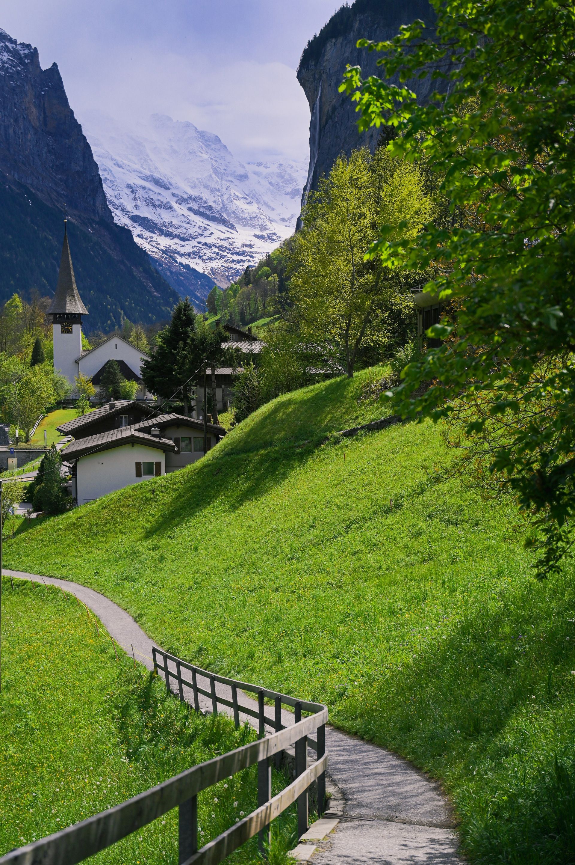 Pathway through lush green hillside towards a village with a church, mountains in the background in Lauterbrunnen.