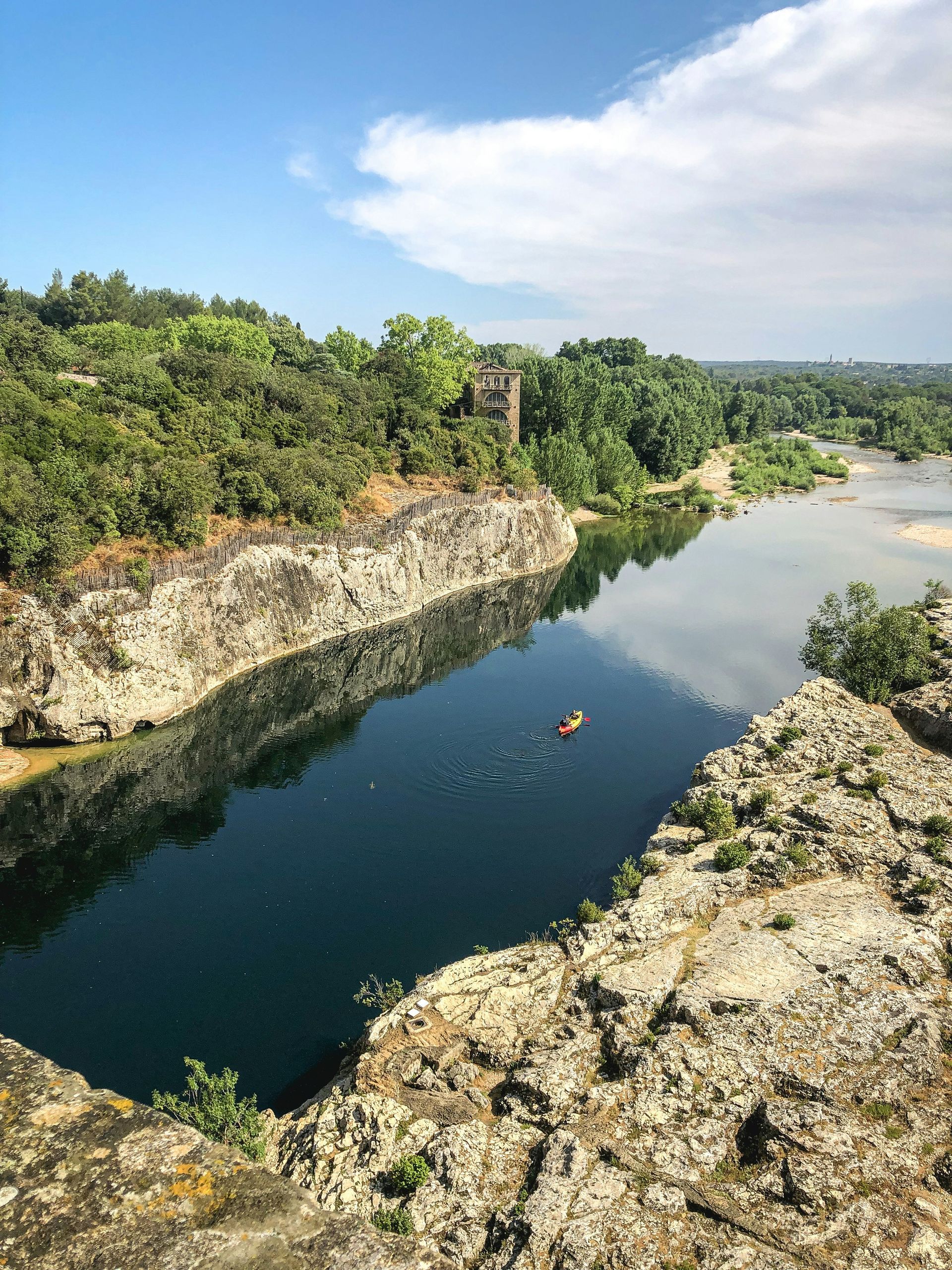 A body of dark water surrounded by rocky cliffs and lush trees, with a distant tower and a person swimming.