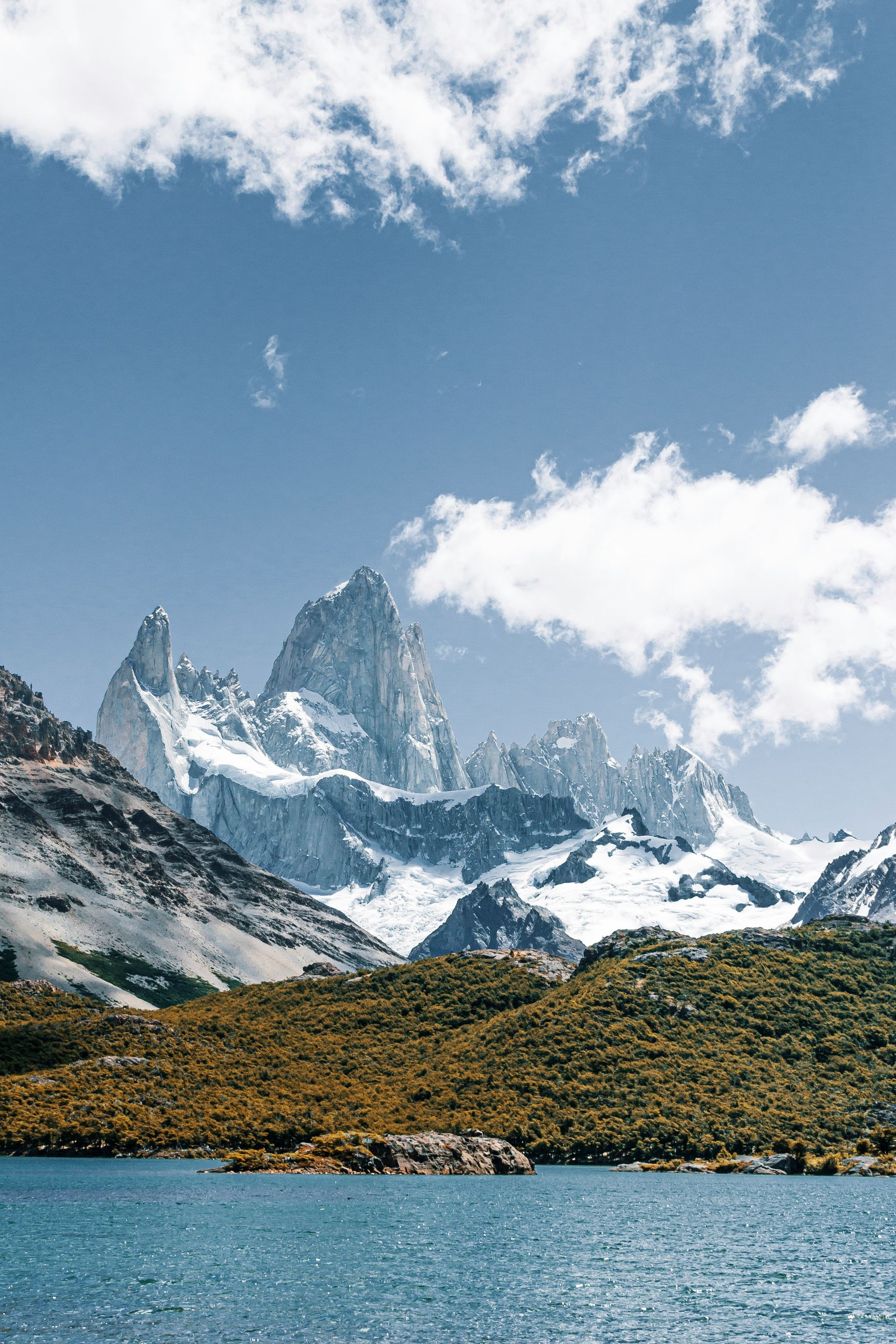 Snow-capped mountains peak above a lake and green shrubs under a blue sky with clouds in El Chaltén, Argentina.