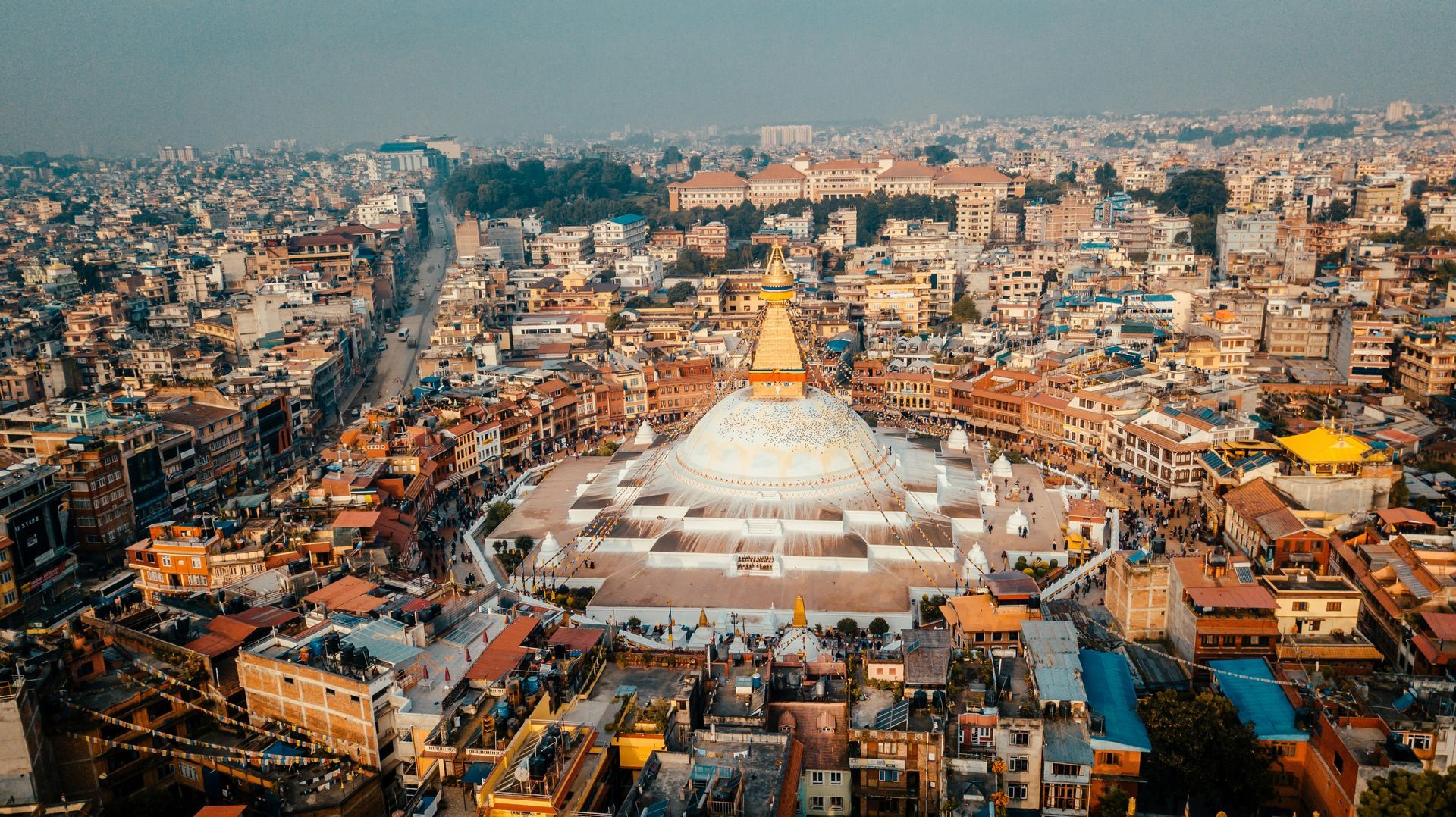 Aerial view of Boudhanath Stupa in Kathmandu, Nepal. White dome surrounded by buildings, a cityscape in soft light.