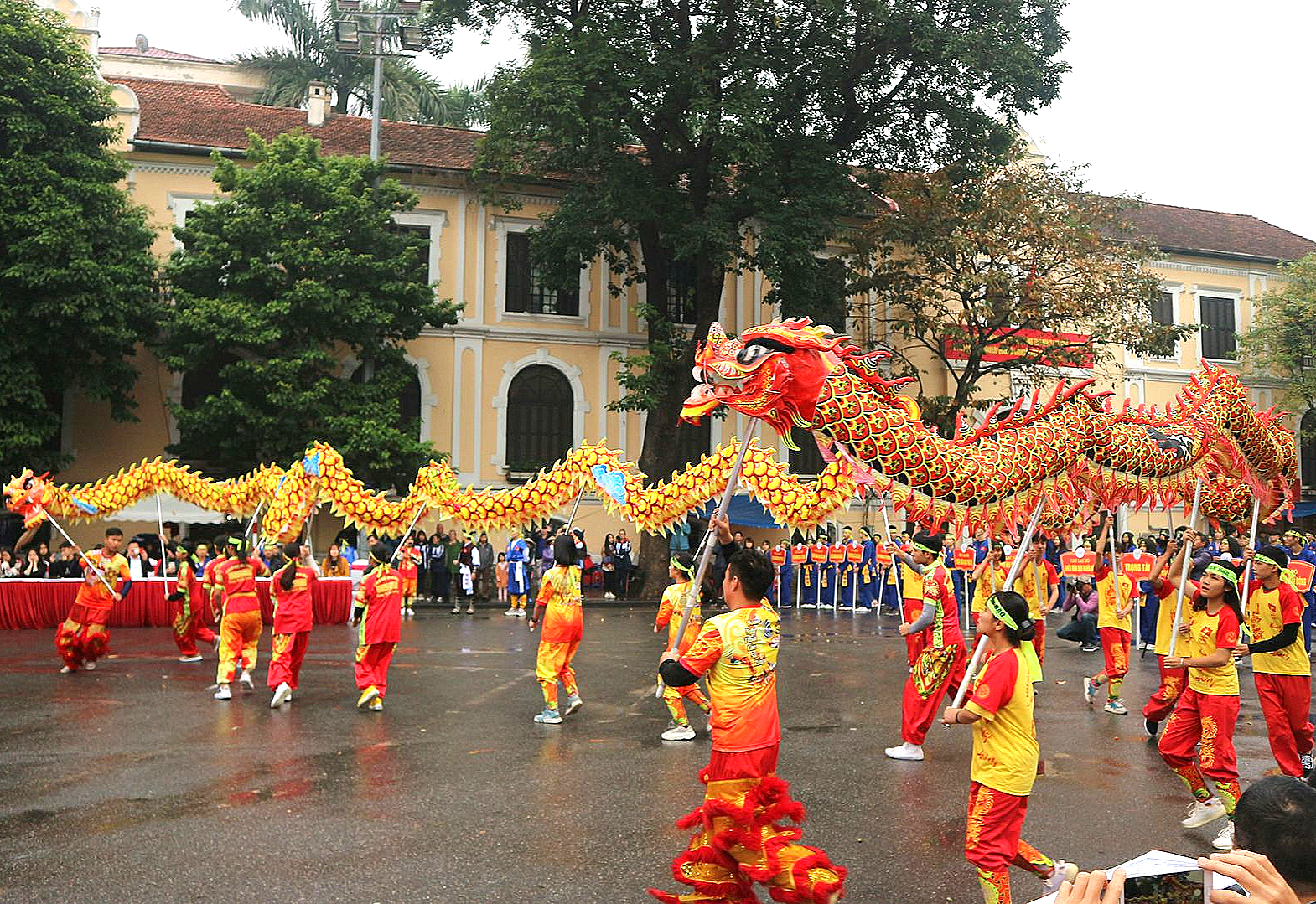 Dragon dance performance in front of a building, red and yellow dragon, people in colorful costumes, outdoors in Vietnam.