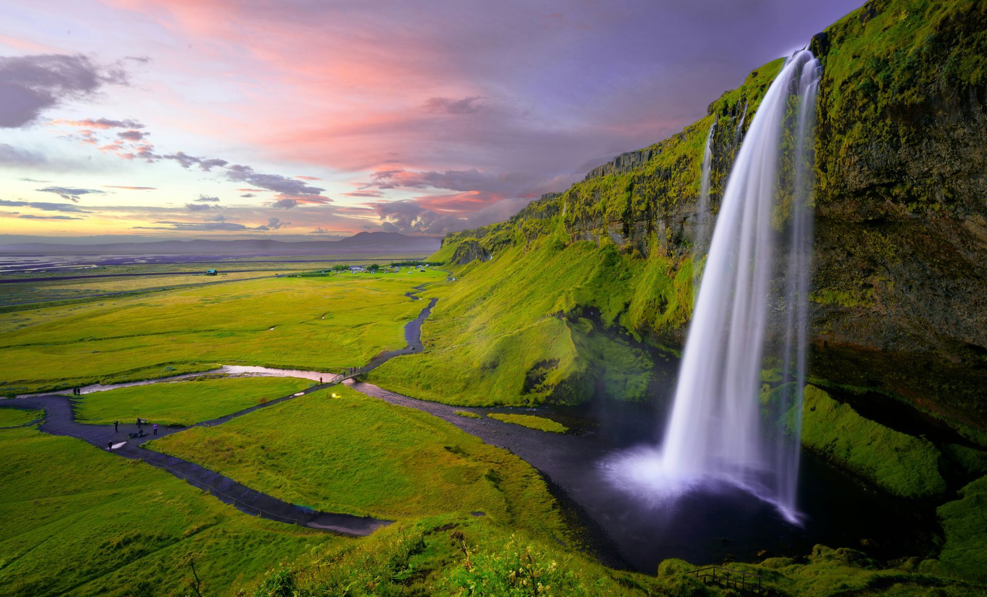 The Seljalandsfoss Waterfall cascading down a green cliffside, flowing into a grassy field with a colorful sunset in Iceland.