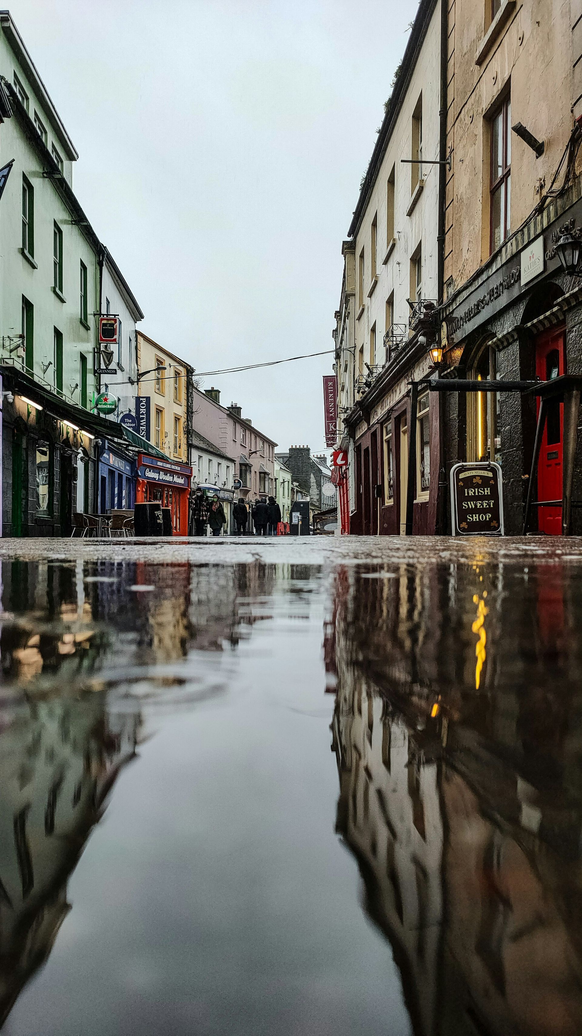 A wet street in Galway, Ireland, reflecting buildings and sky; pedestrians in the distance.