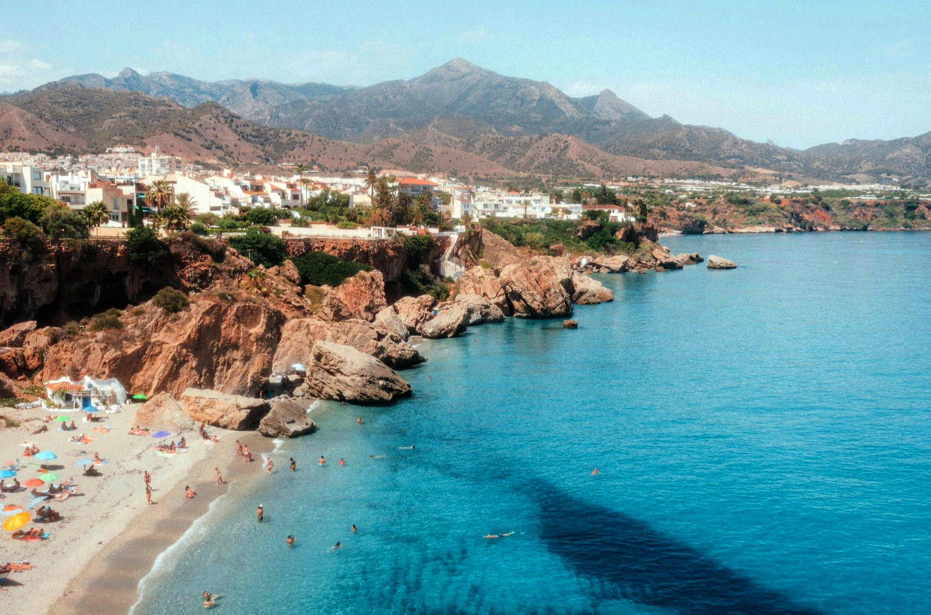 Beach with clear turquoise water, rocky cliffs, and a town with white buildings at the mountains' base in Spain.