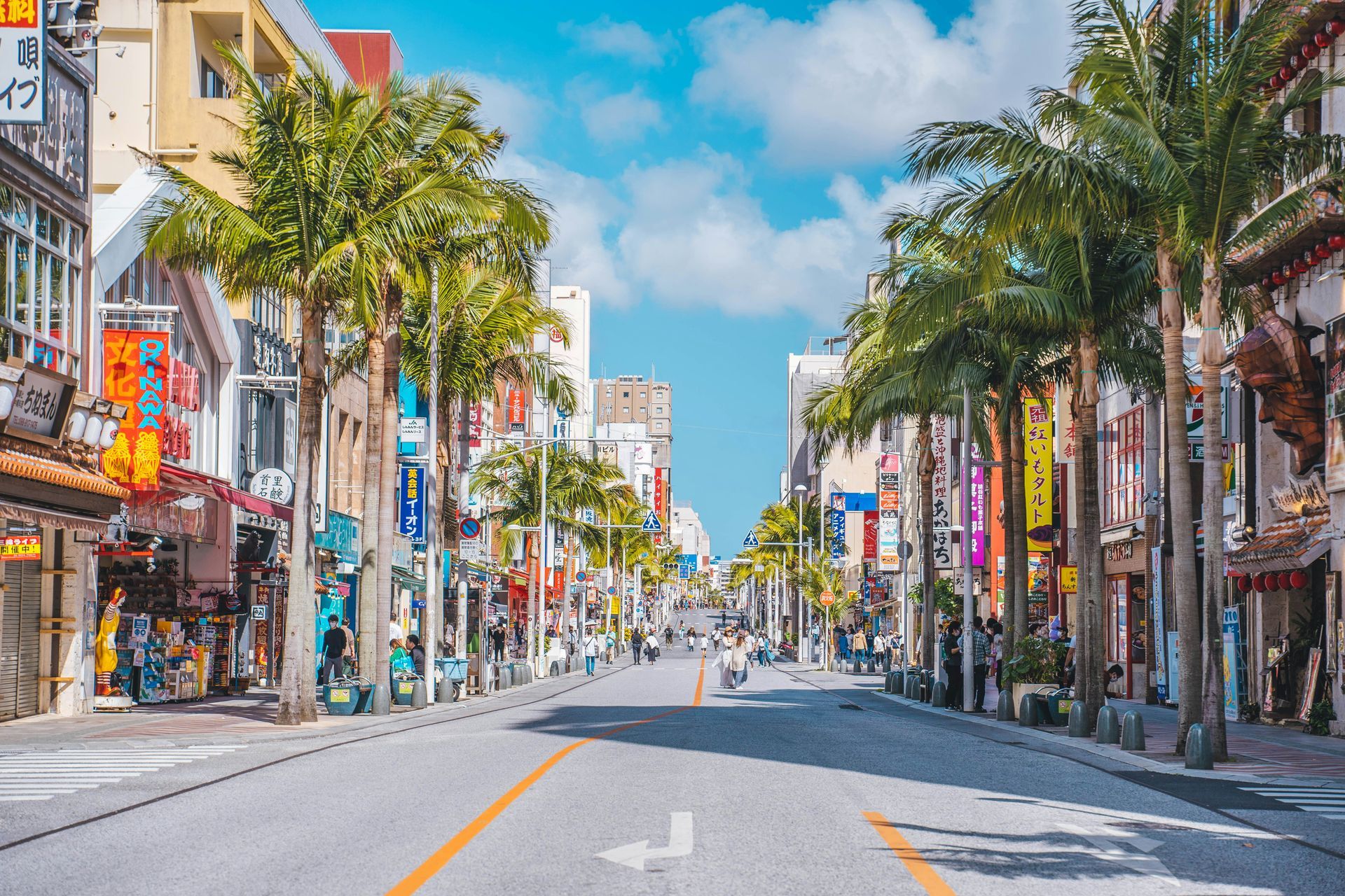 Street lined with shops and palm trees in Okinawa, Japan. Bright sky, people walking.