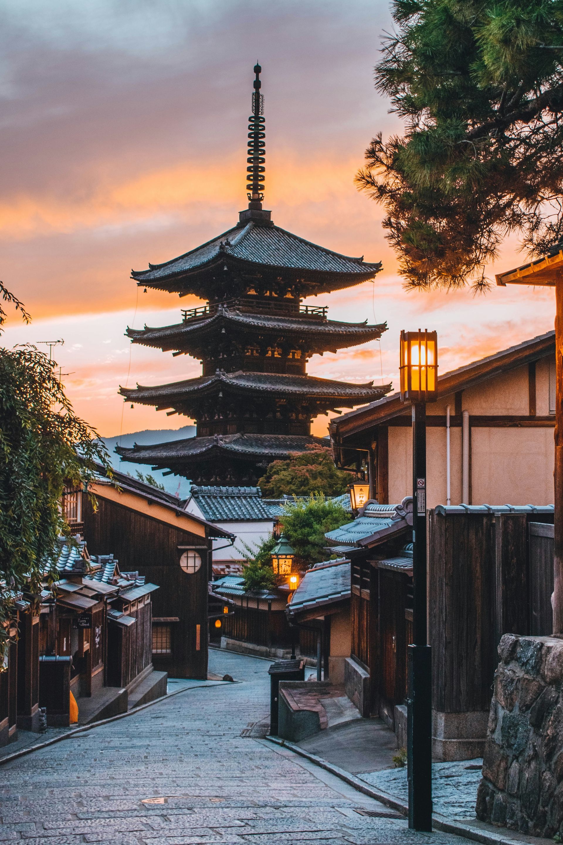 Kyoto street scene with five-story pagoda at sunset; stone path, wooden buildings, and orange sky.