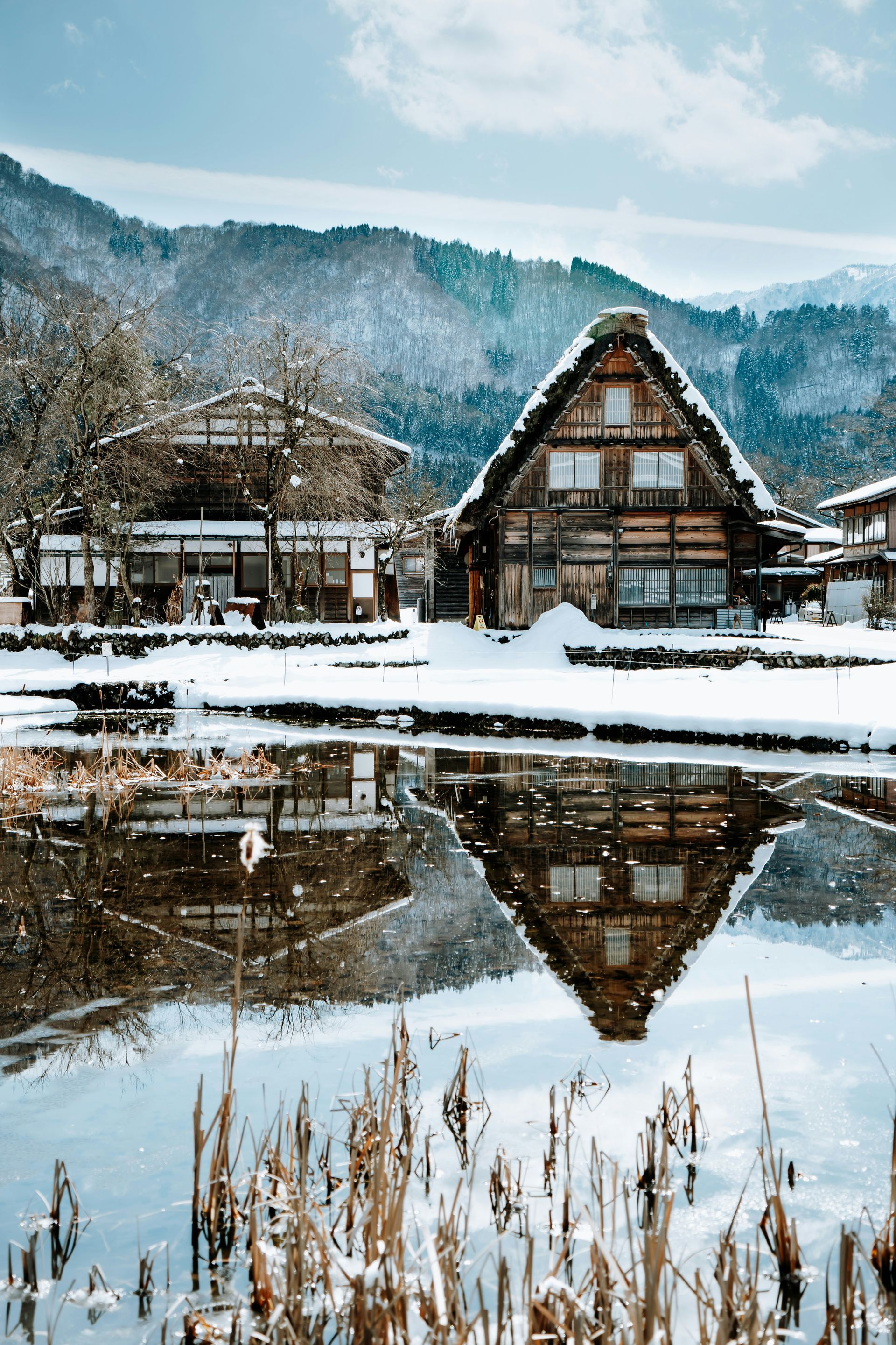 Snow-covered traditional Japanese thatched-roofed houses on a pond; snowy mountains in the background in Shirakawa, Japan.