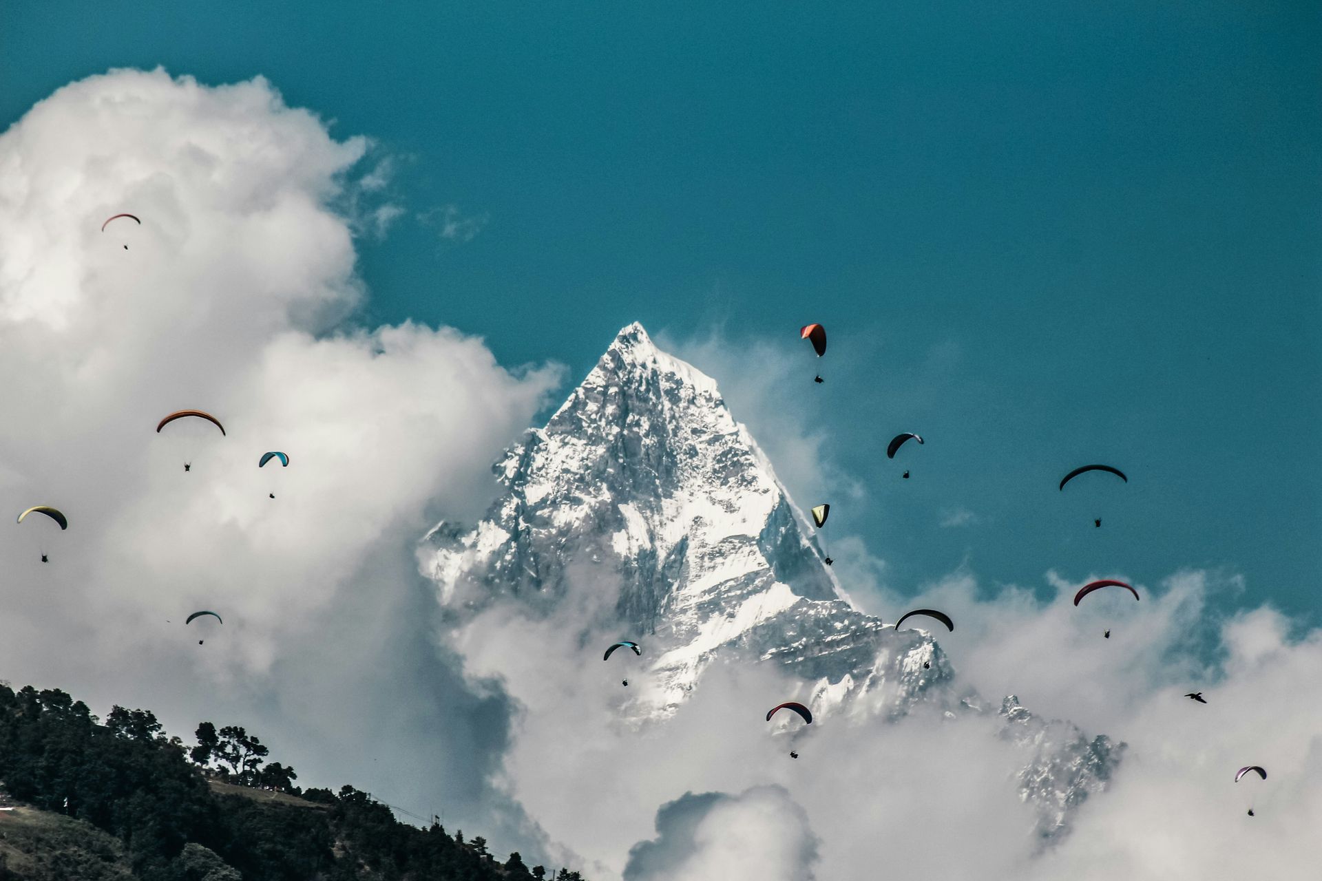 Paragliders soar near a snowy mountain peak, amidst fluffy clouds in Pokhara, Nepal.