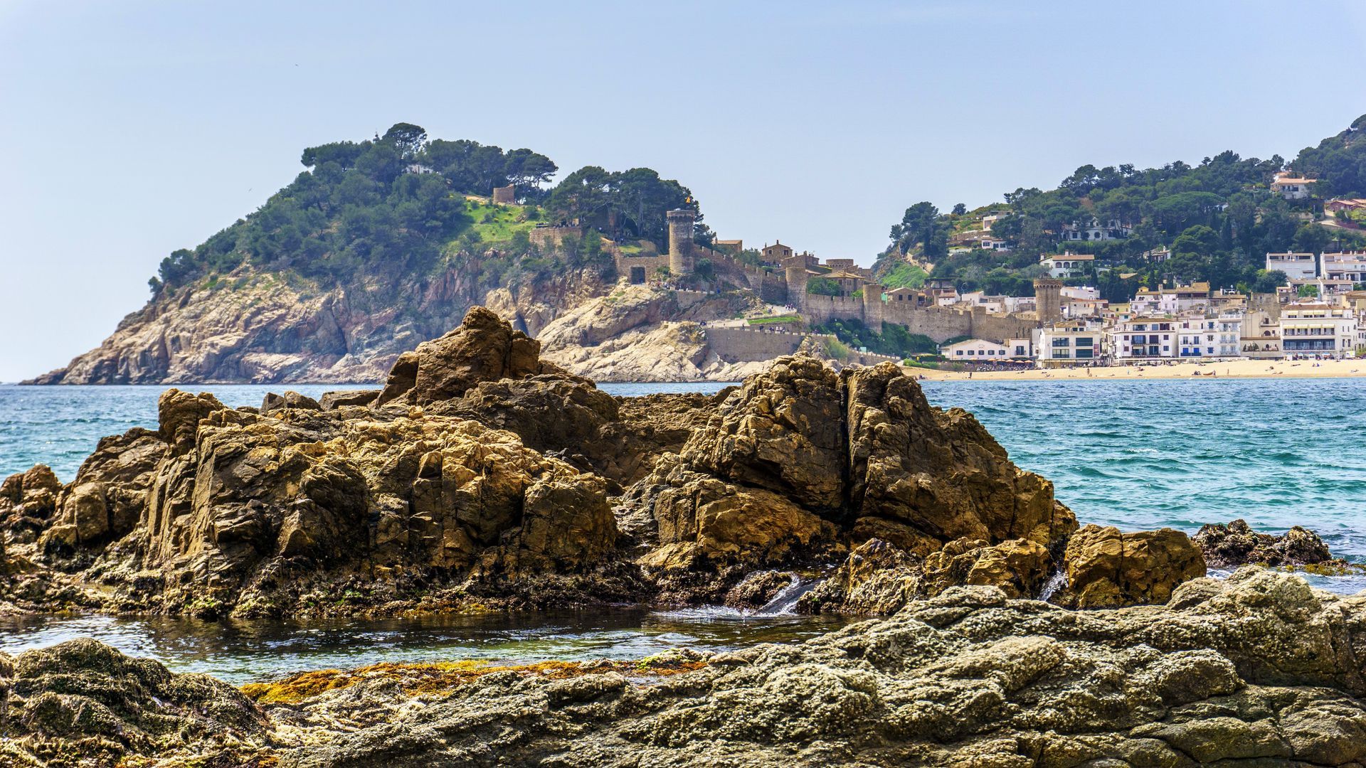 A large rock in the middle of the ocean with Tossa de Mar, Spain in the background.