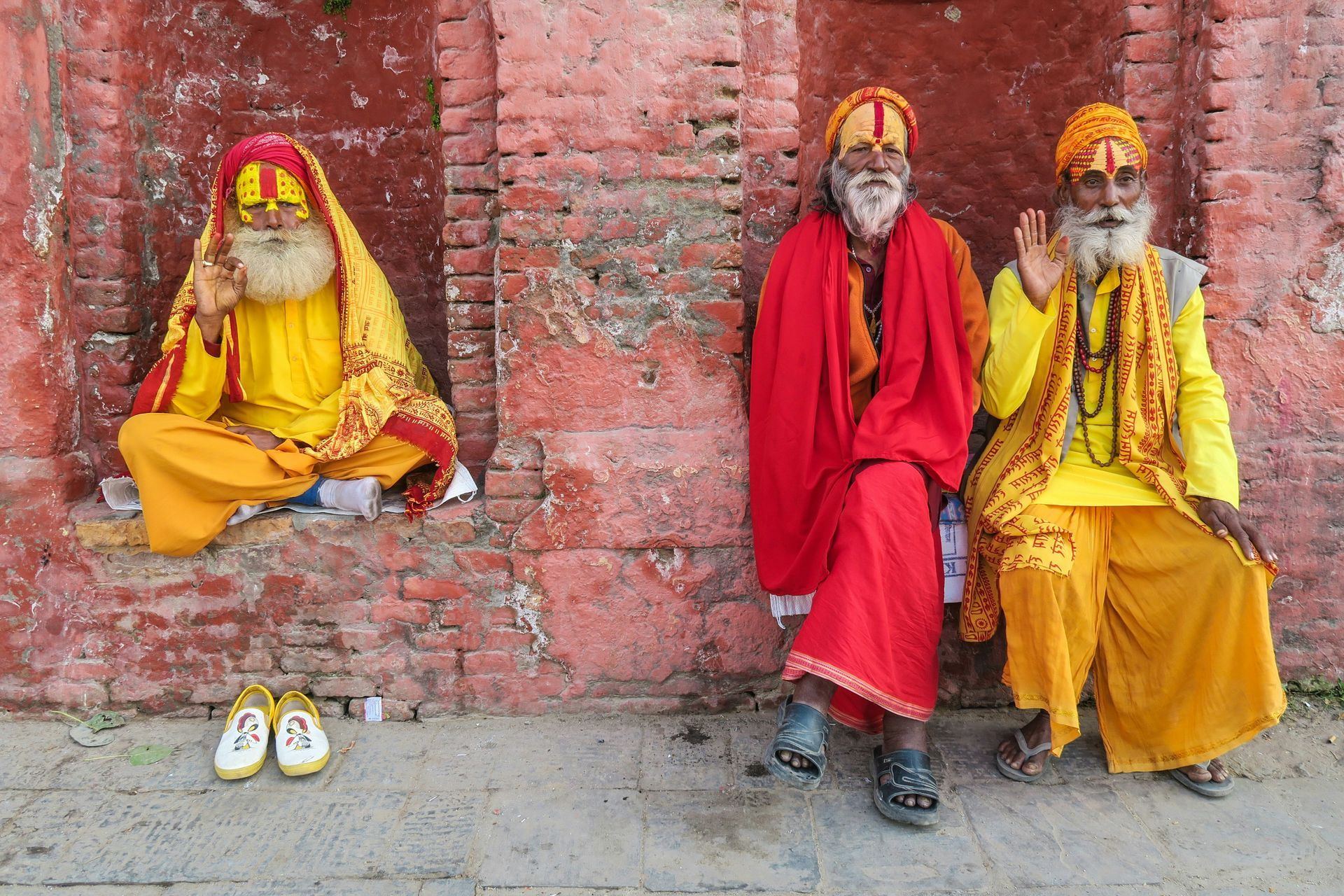 Three robed Nepalese men with long beards and headwear seated against a red brick wall, one with hand raised, shoes nearby. 