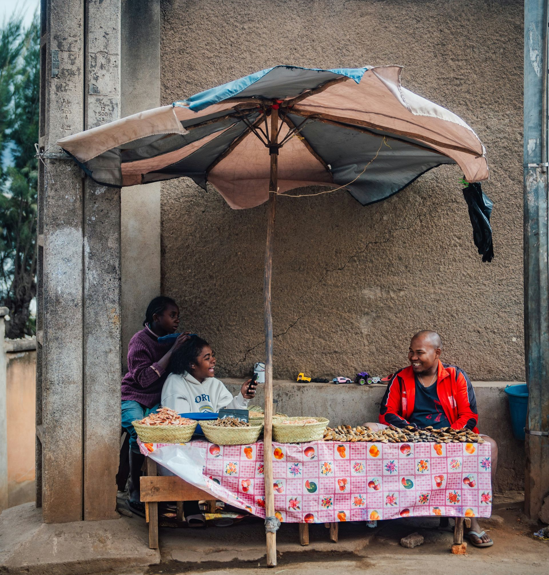 People at a street food stall beneath an umbrella. One braids hair in Madagascar.