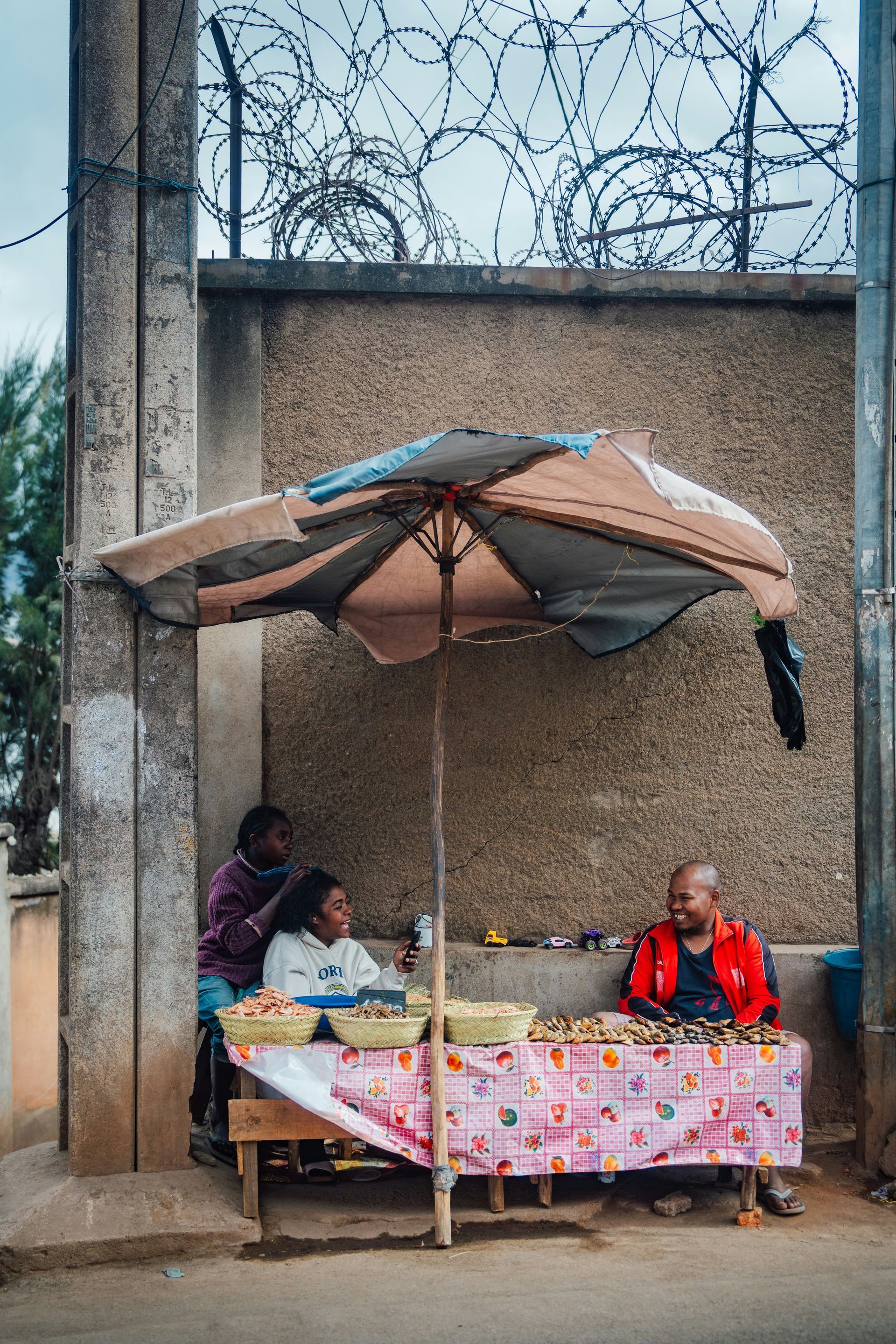 People at a street food stall beneath an umbrella. One braids hair in Madagascar.