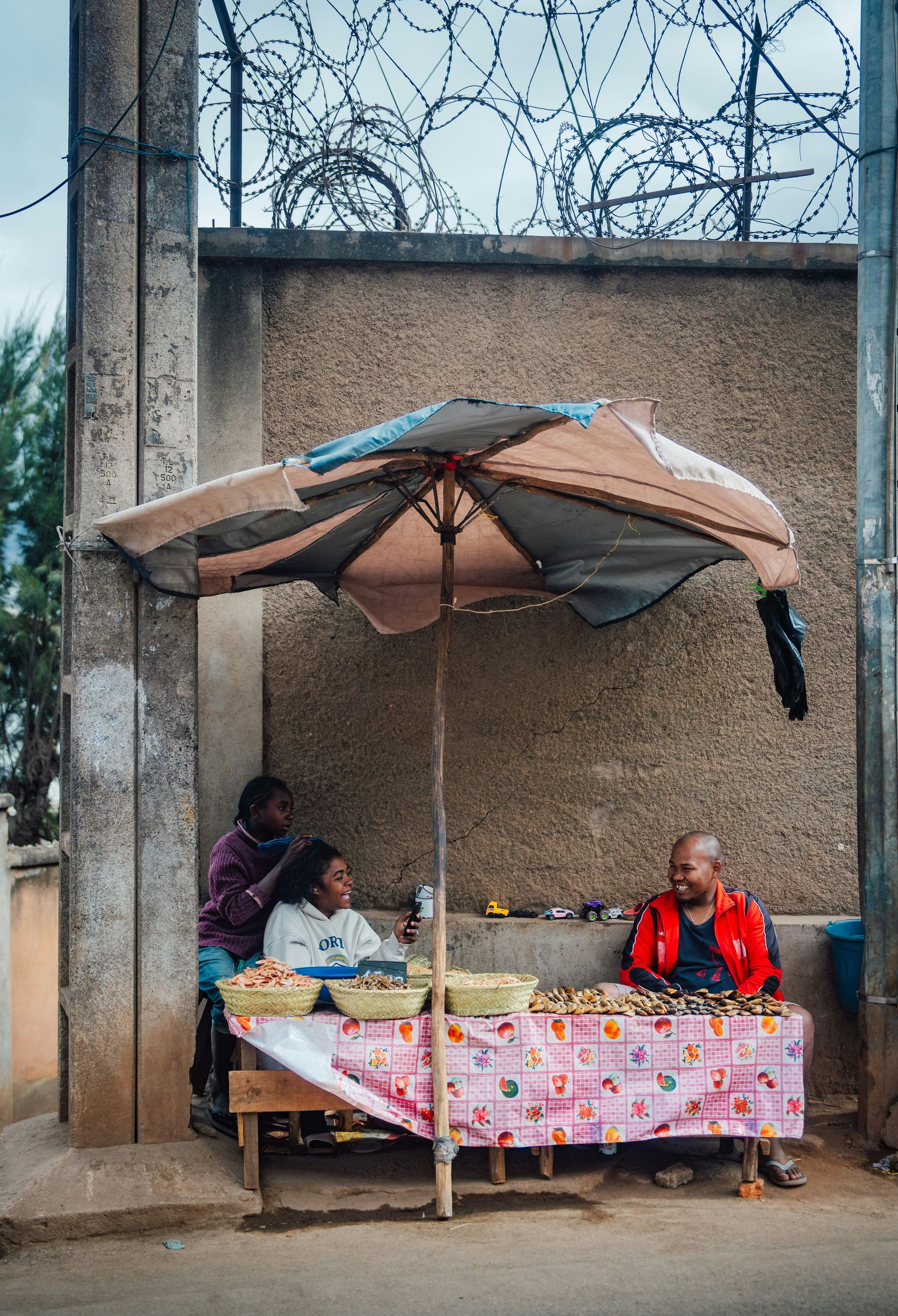 People at a street food stall beneath an umbrella. One braids hair in Madagascar.