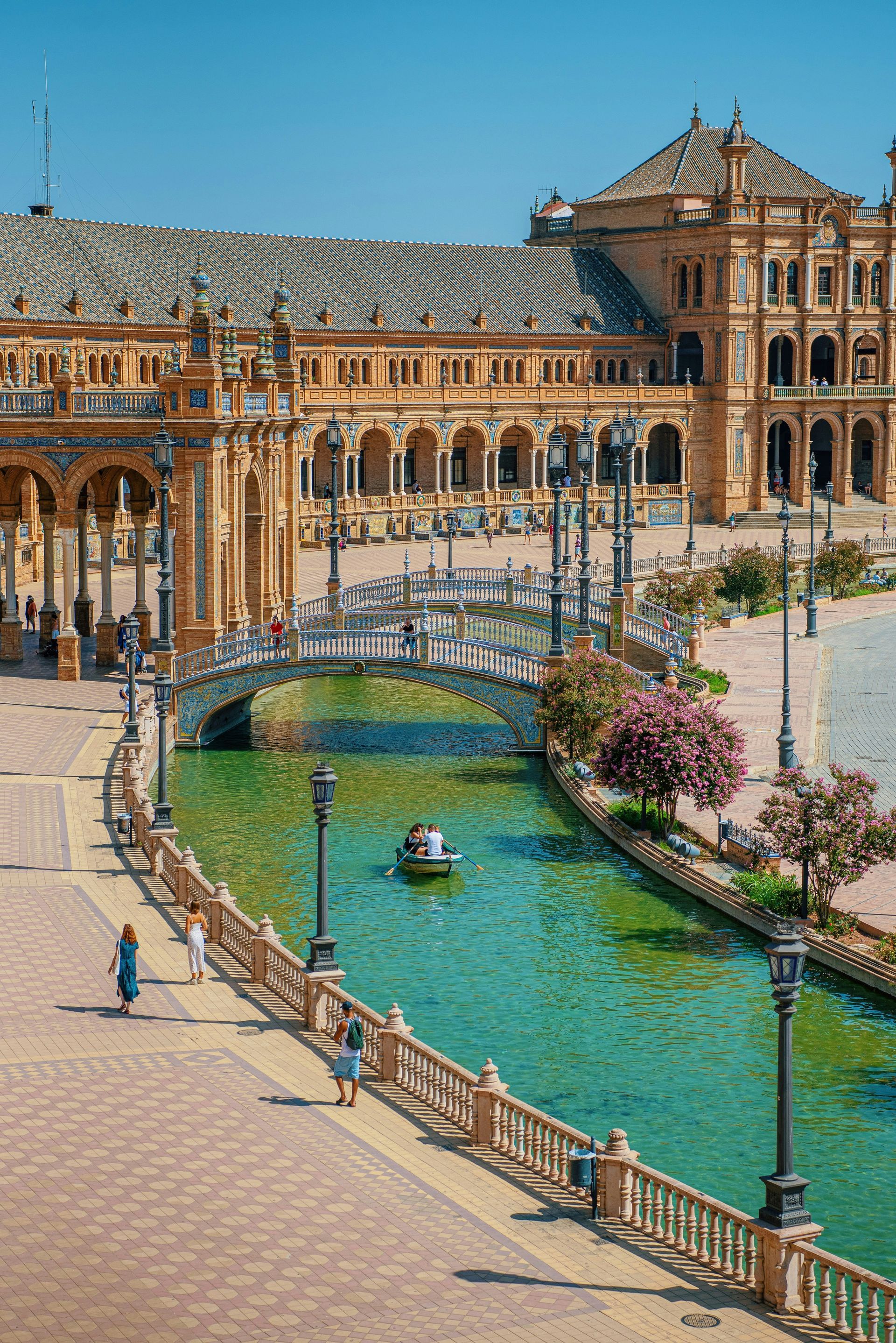 Plaza de España in Seville, Spain: ornate buildings surround a canal with a bridge and boaters; sunny, blue sky.