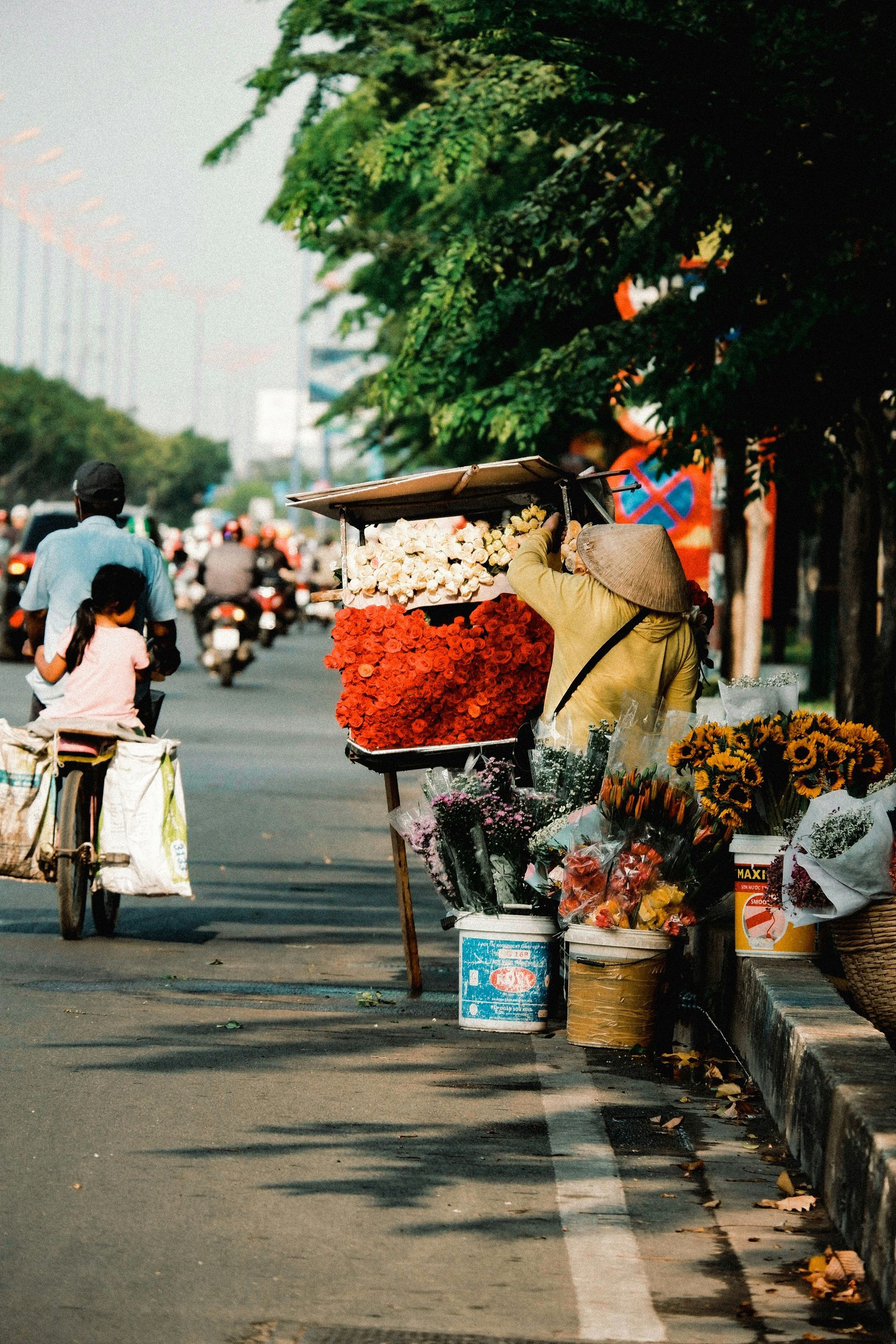 Flower stand on a city street with colorful blooms. A cyclist and motorbike riders pass by in Ho Chi Minh, Vietnam.