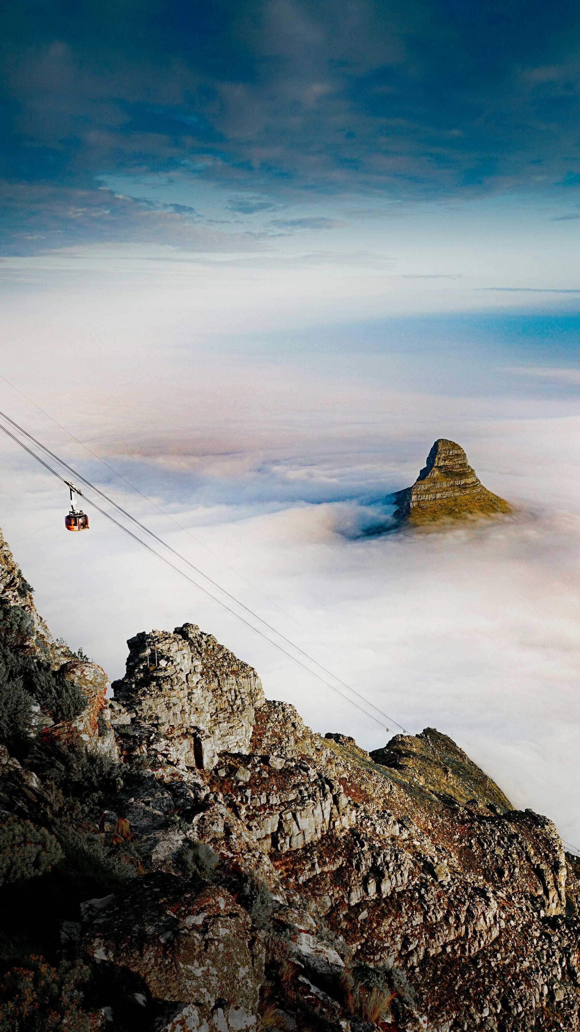 Cable car ascending from rocky mountain into a sea of clouds, another peak visible in Cape Town, South Africa.