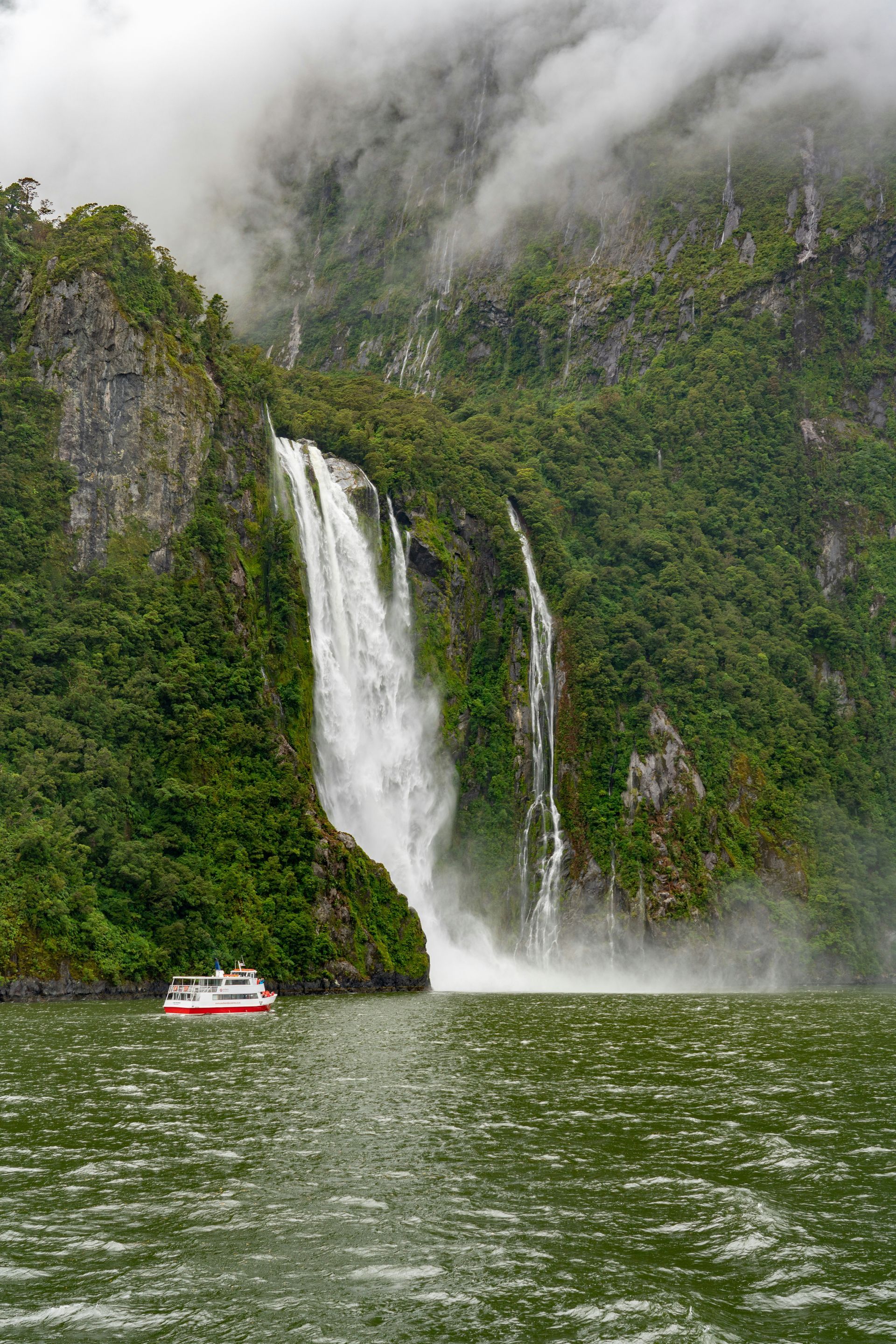 A boat is floating in front of a waterfall in the middle of the Milford Sound in Fiordland National Park, New Zealand.