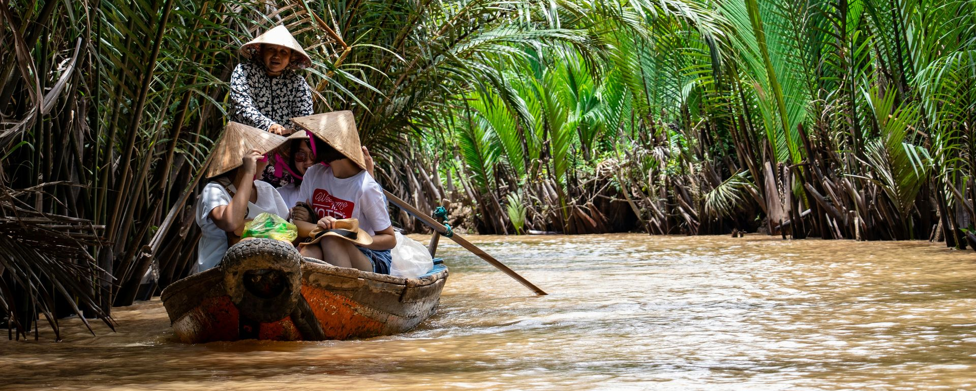 People in conical hats ride a boat on a the Mekong River surrounded by lush green plants.