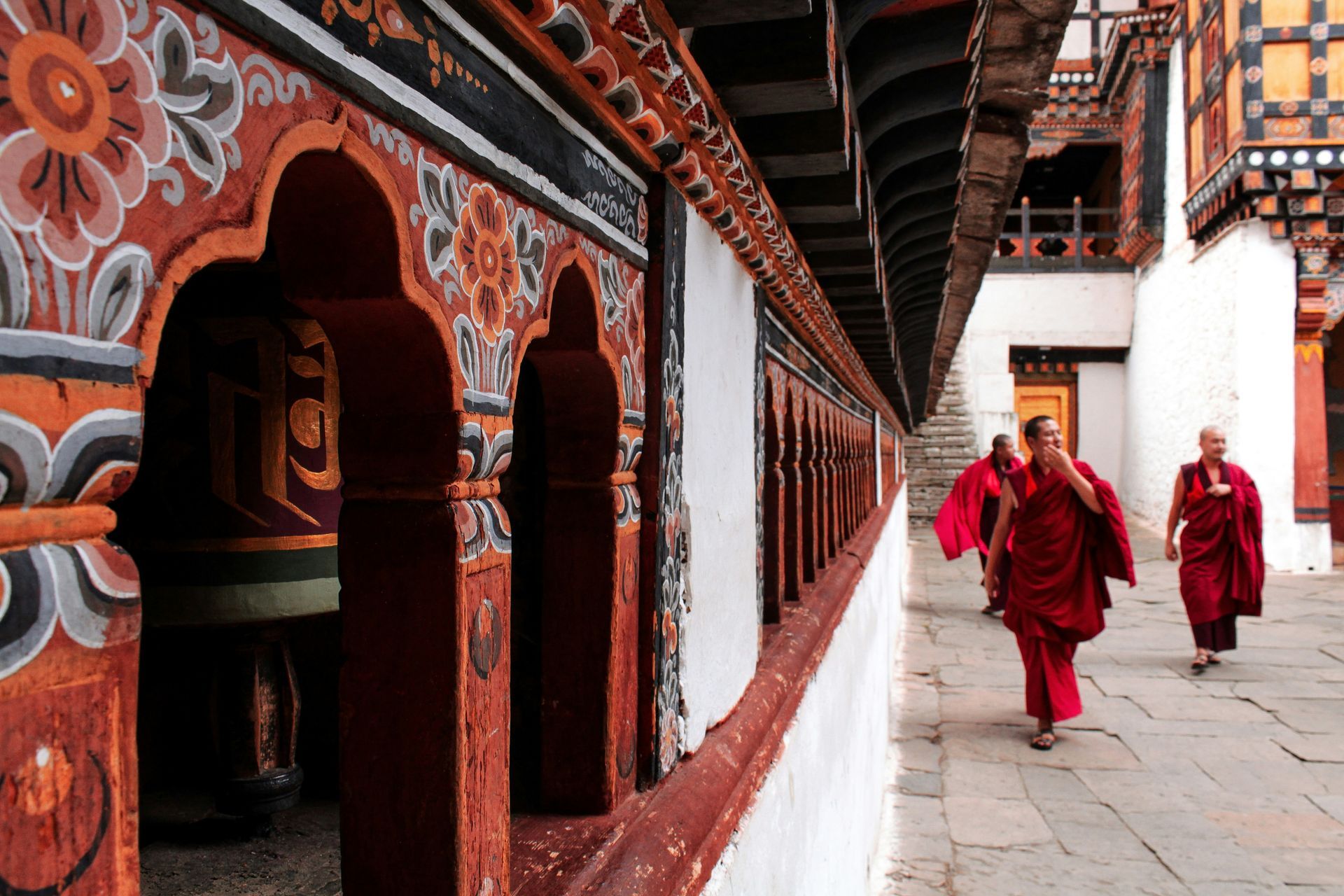 Monks in red robes walk along a corridor, intricate orange and white patterns adorn walls in Paro, Bhutan.