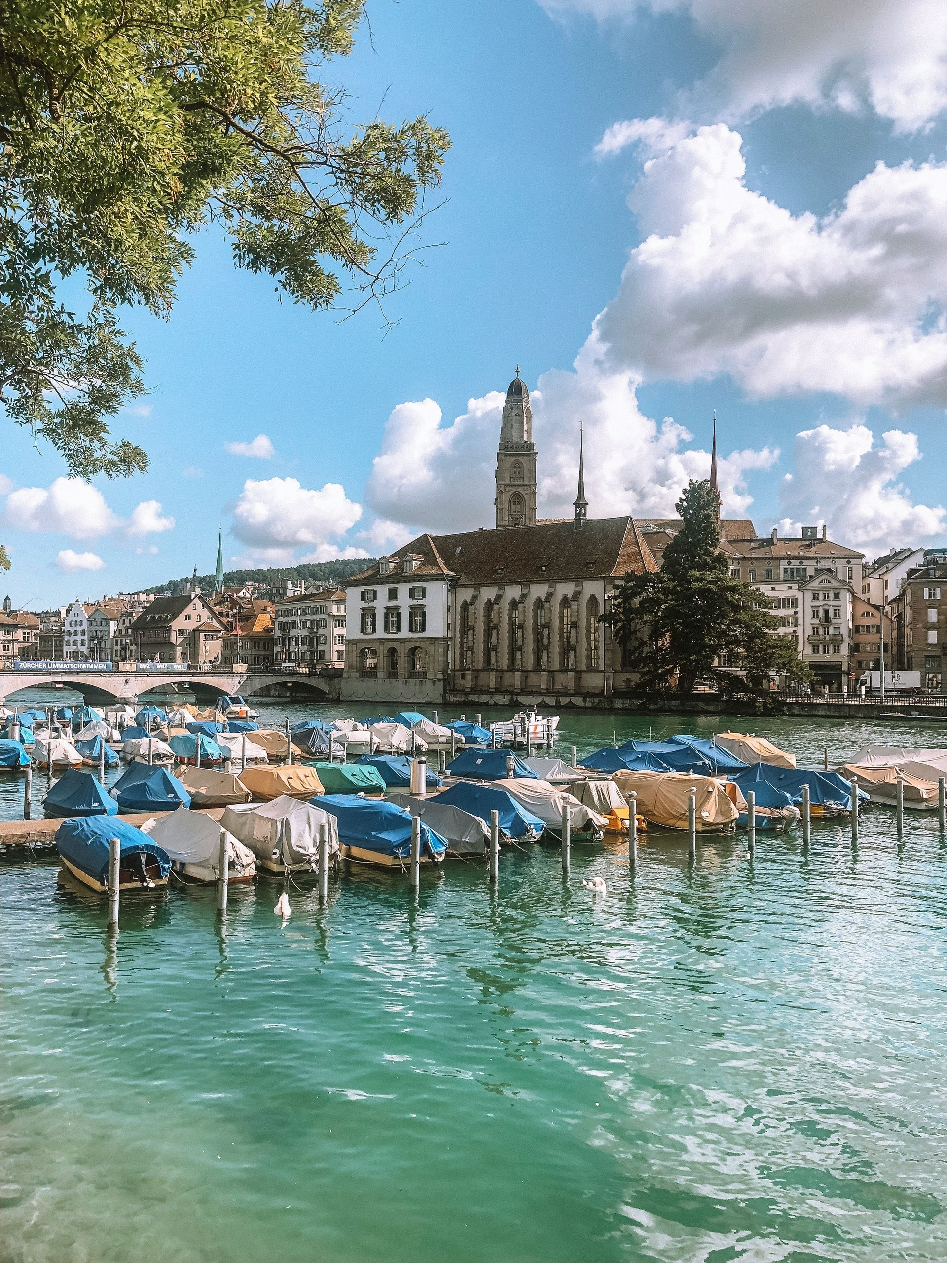 Boats docked on turquoise water, Zurich, Switzerland.  Buildings line the shore under a cloudy blue sky.
