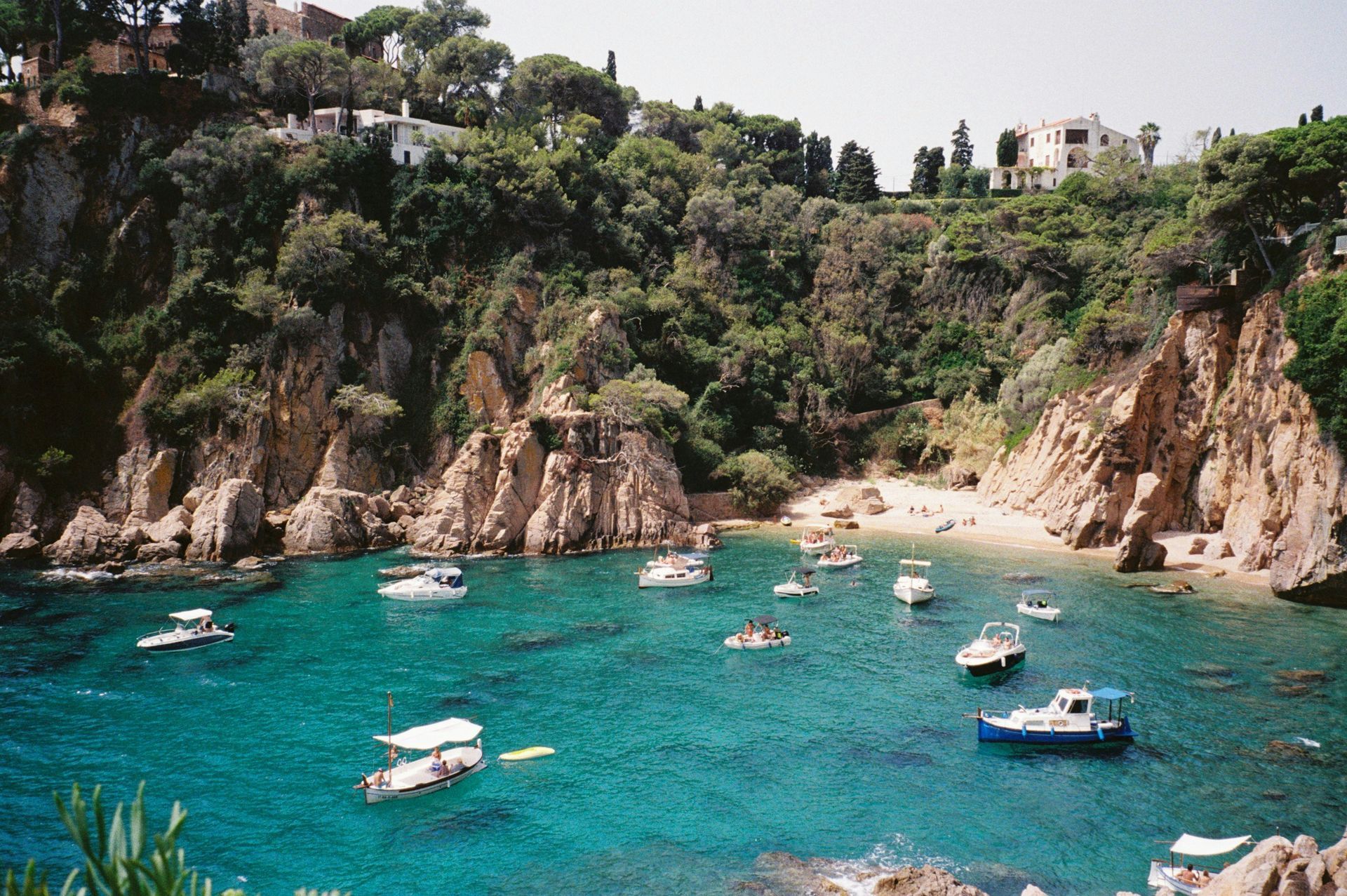 Boats in a turquoise cove nestled between rocky cliffs and lush greenery on Costa Brava, Spain.