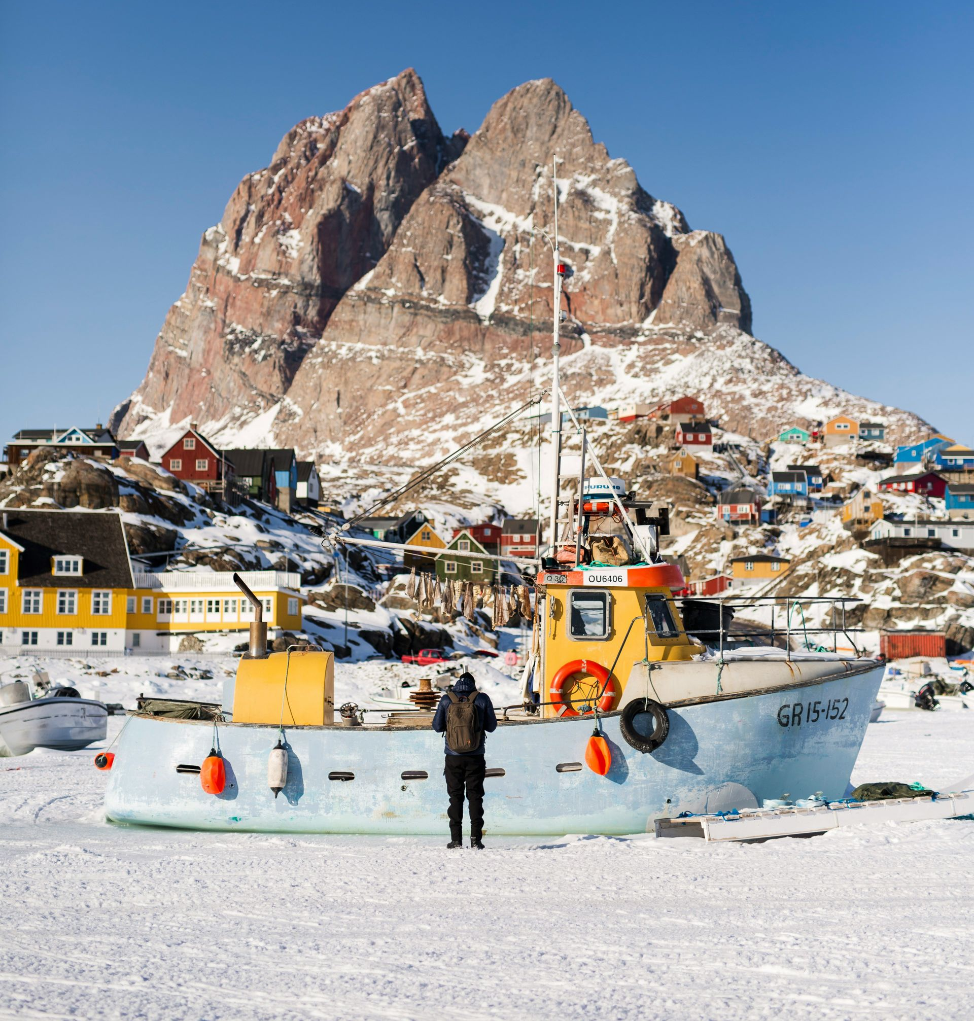 Person standing by a boat, snowy landscape, village with colorful houses, mountain backdrop in Uummannaq, Greenland.