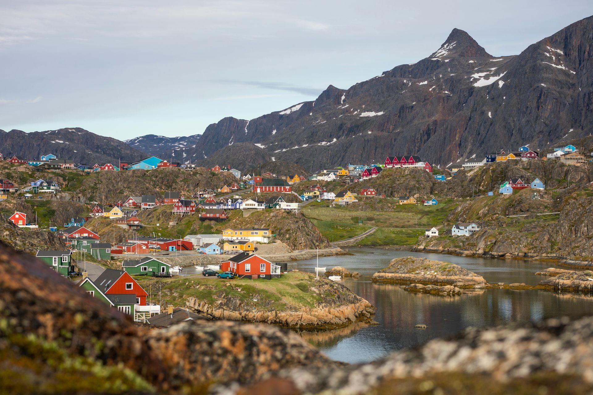 Colorful houses dot a rocky landscape in Sisimiut, Greenland, in front of mountains and calm water in the foreground.