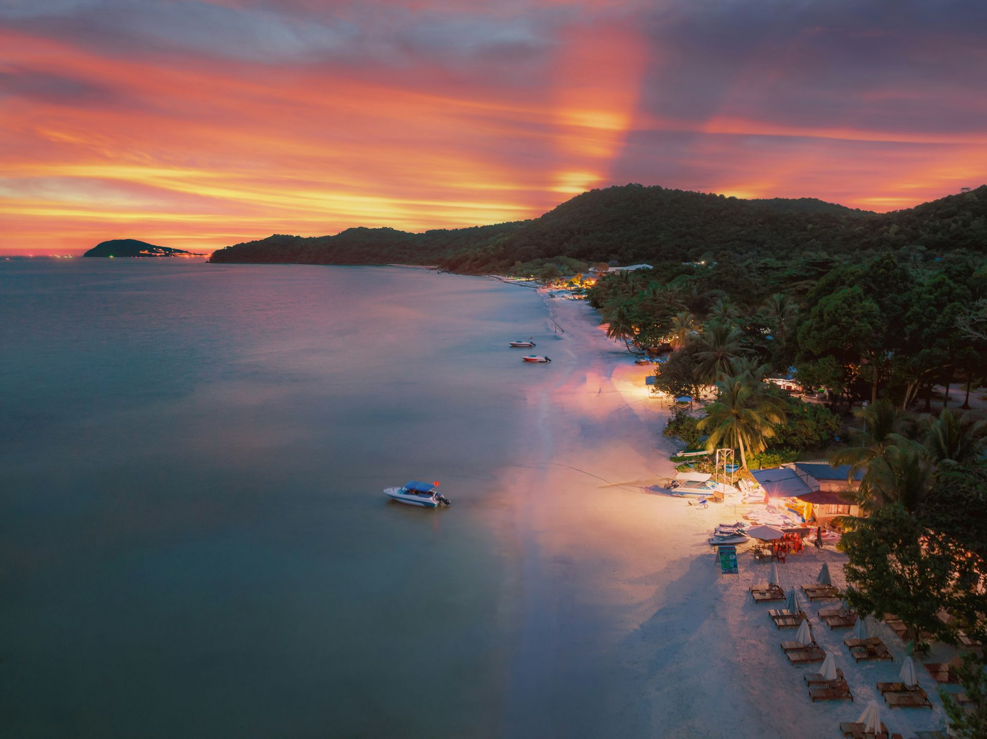 Sunset over a beach with boats. Orange and pink sky, calm water, and lights on the shore in Phú Quốc, Vietnam.