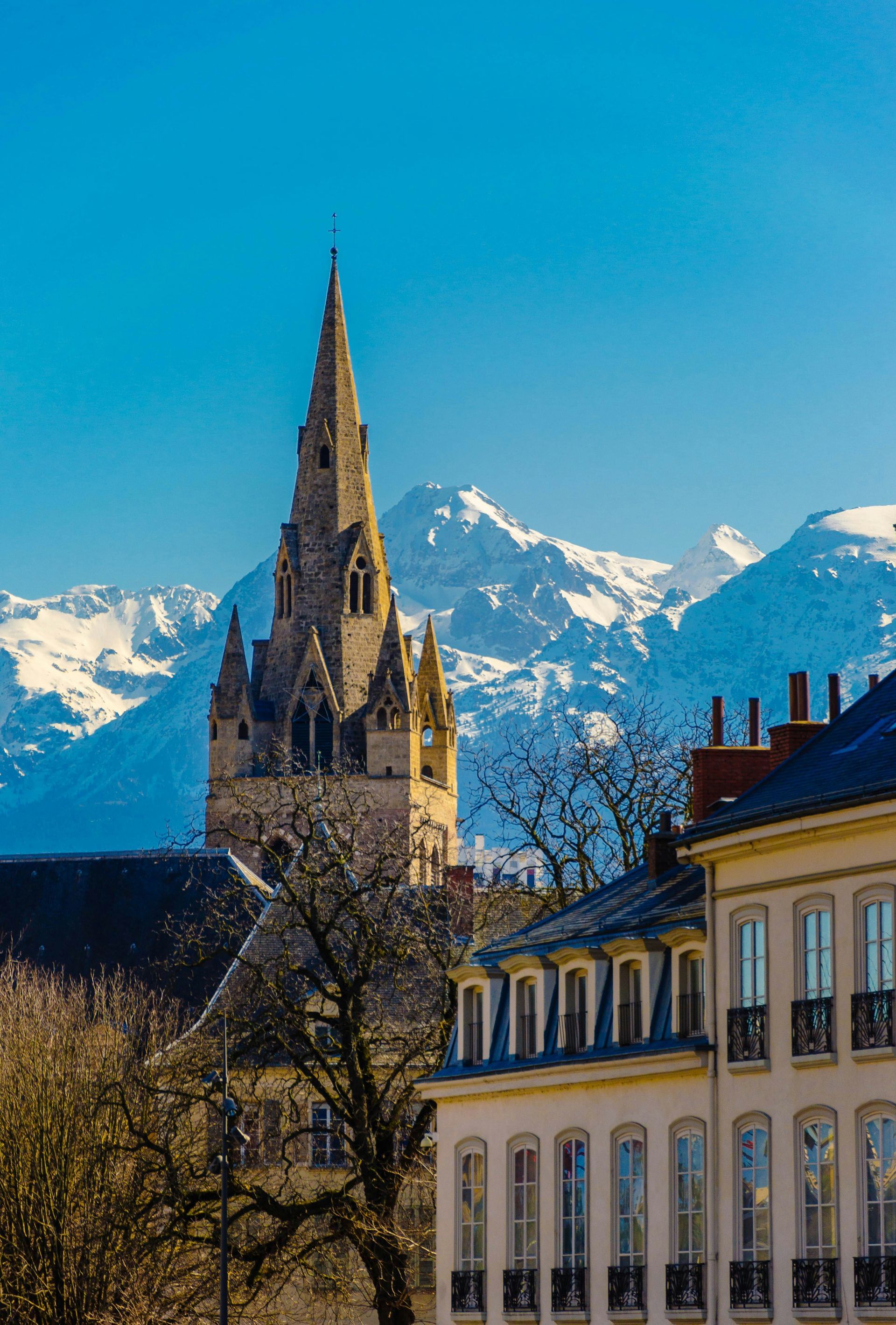 A church with a steeple in the middle of a city with snowy mountains in the background in Grenoble, France.
