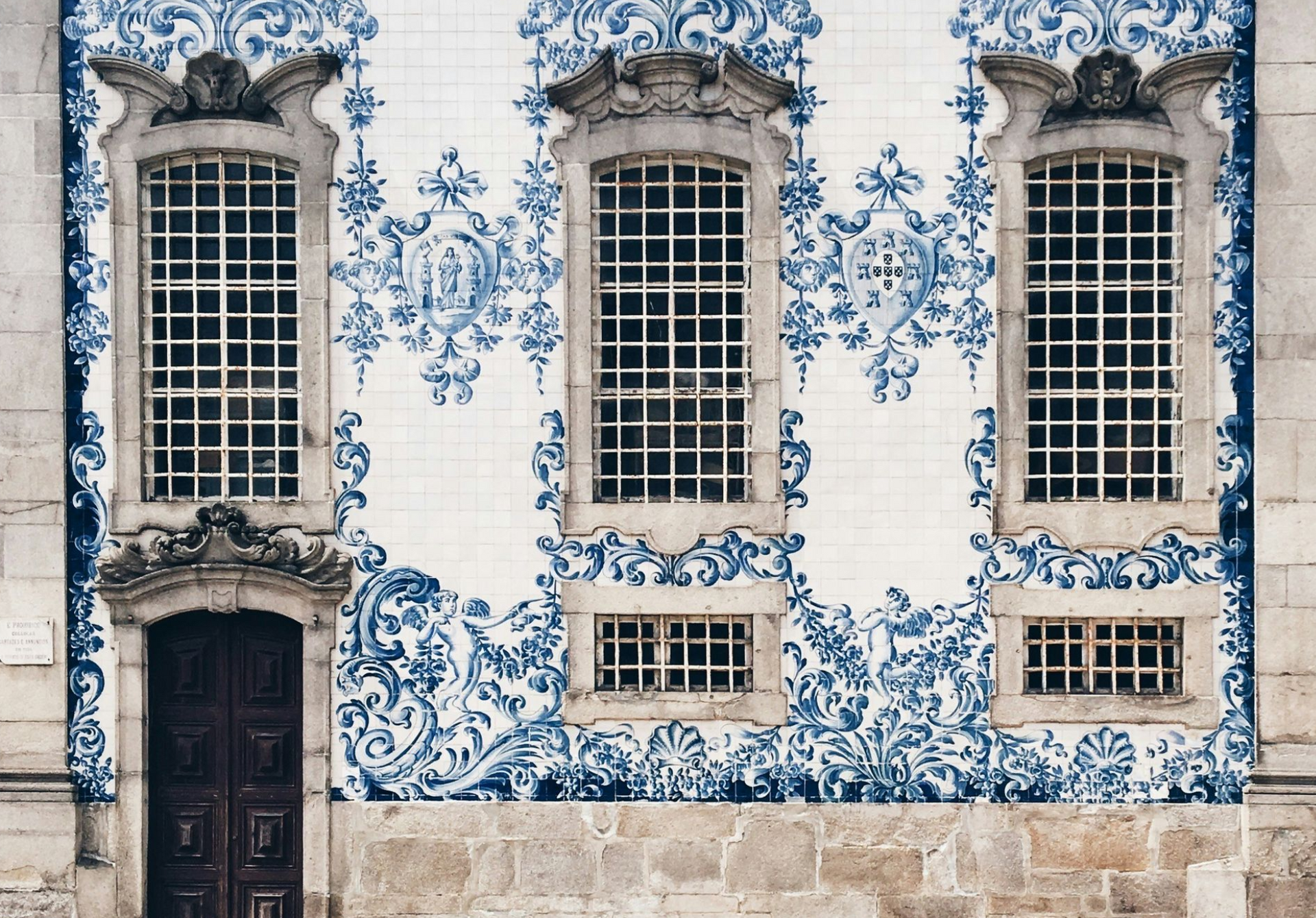 Exterior of a building with blue and white azulejos tile, ornate windows, and a dark wooden door in Porto, Portugal.