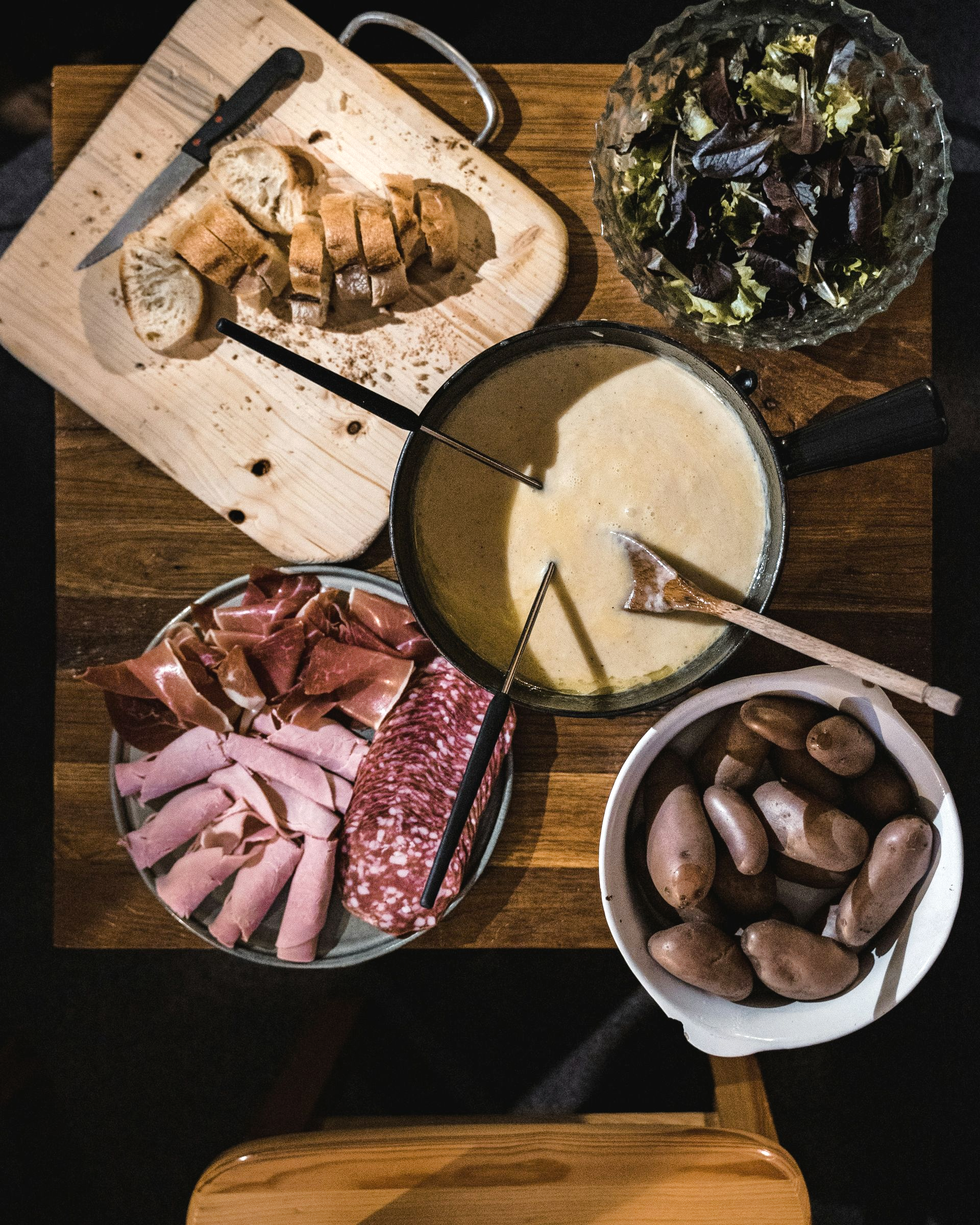 Fondue meal: cheese pot, charcuterie, bread, potatoes, salad, on wood table in Switzerland.