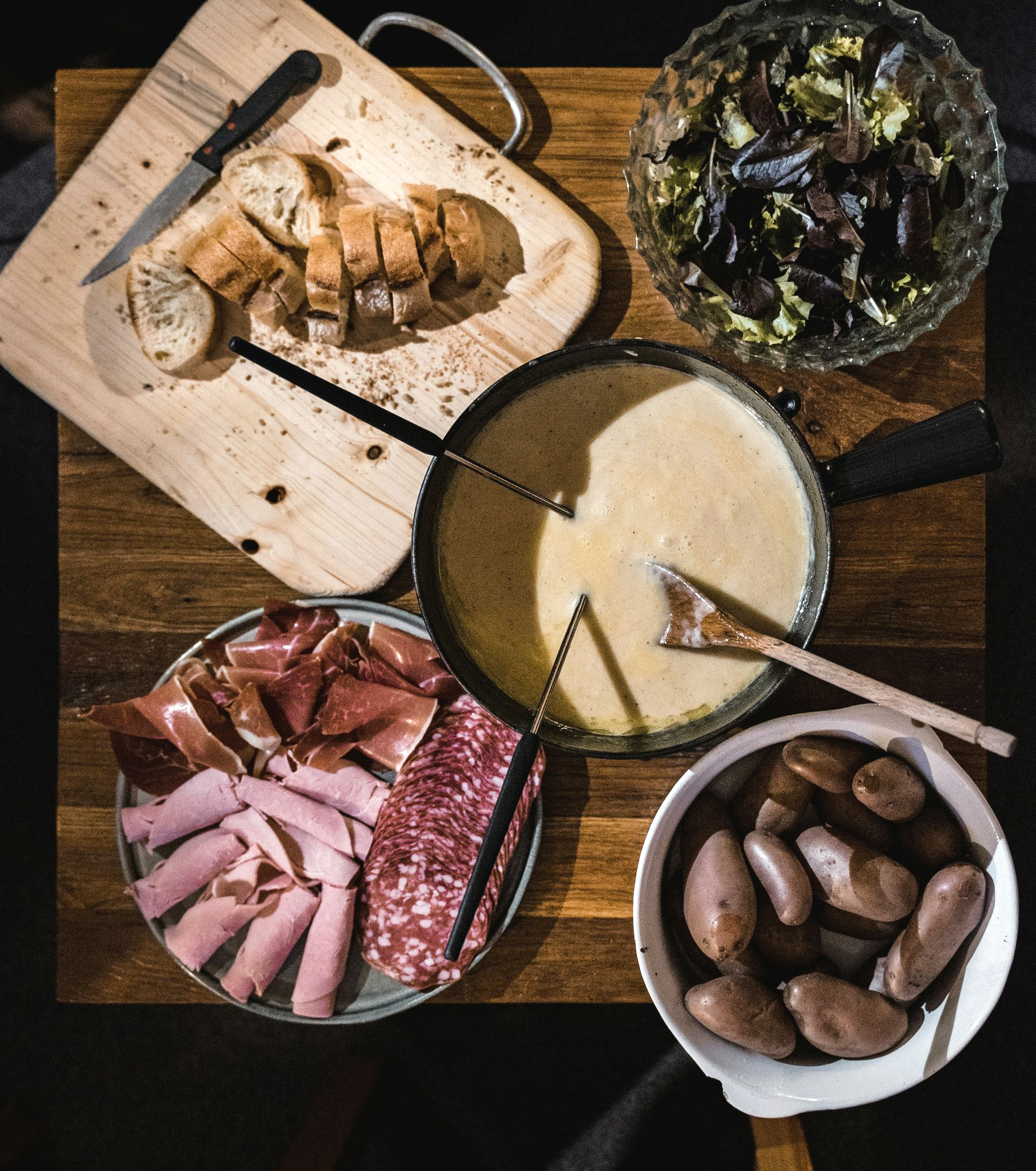 Fondue meal: cheese pot, charcuterie, bread, potatoes, salad, on wood table in Switzerland.