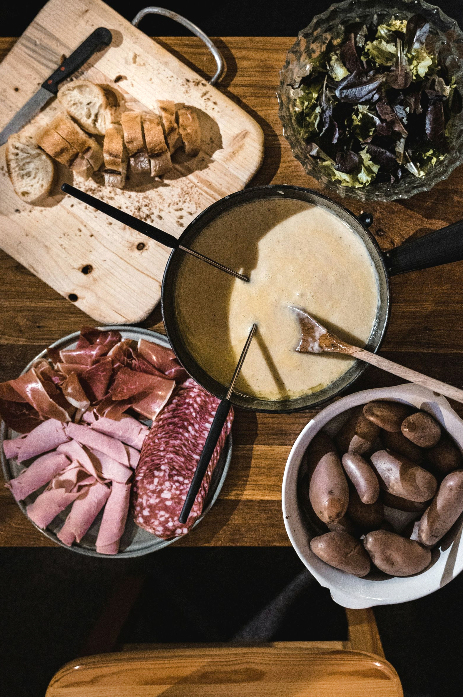 Fondue meal: cheese pot, charcuterie, bread, potatoes, salad, on wood table in Switzerland.