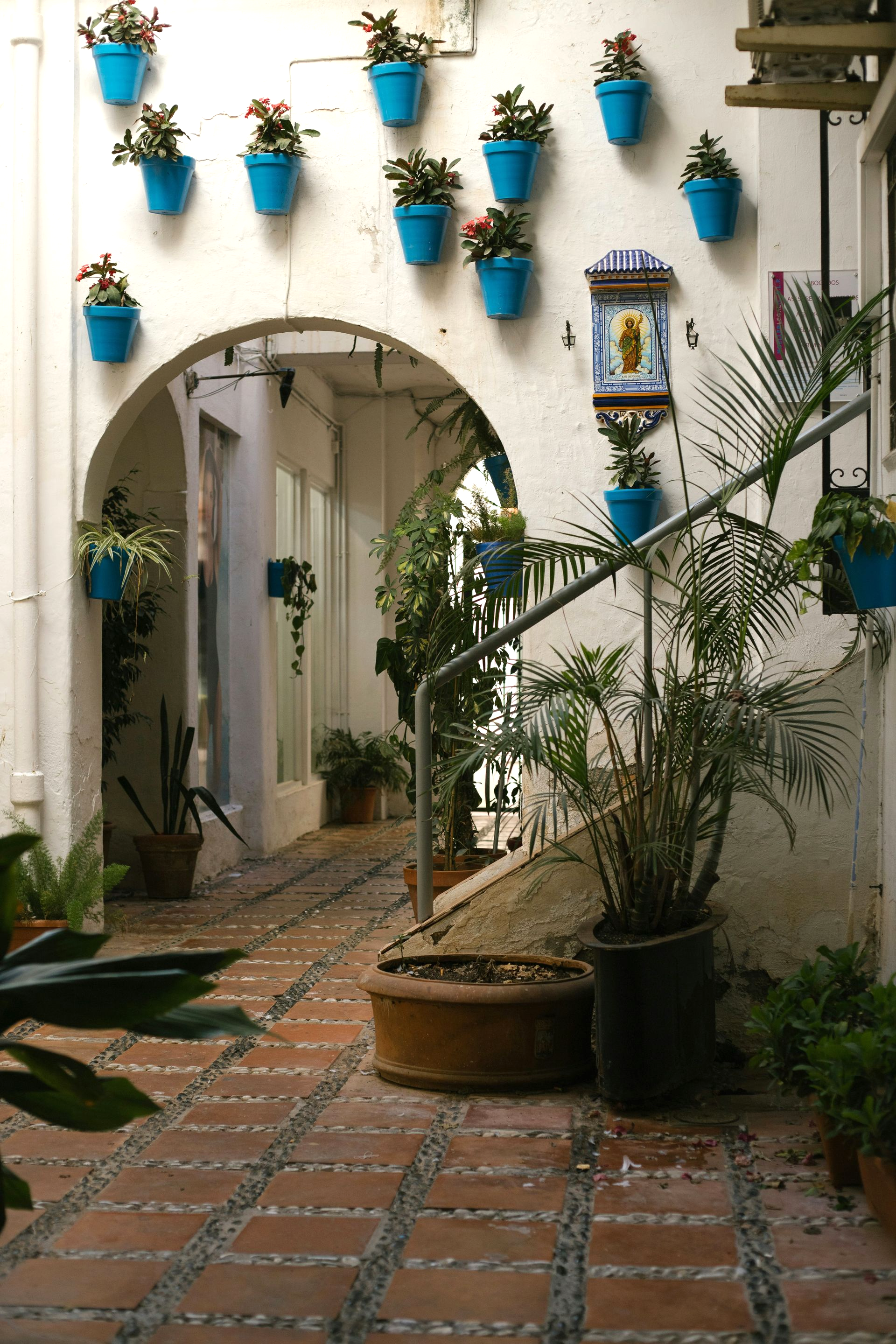 Whitewashed alley with archway, vibrant blue flower pots, brick path, lush greenery in Marbella, Spain.