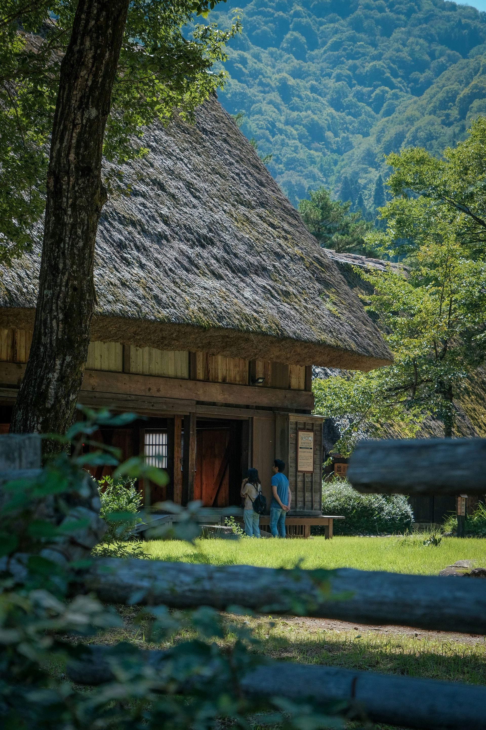 Two figures in front of a wooden cottage with a thatched roof, mountains in the background in Shirakawa, Japan. 