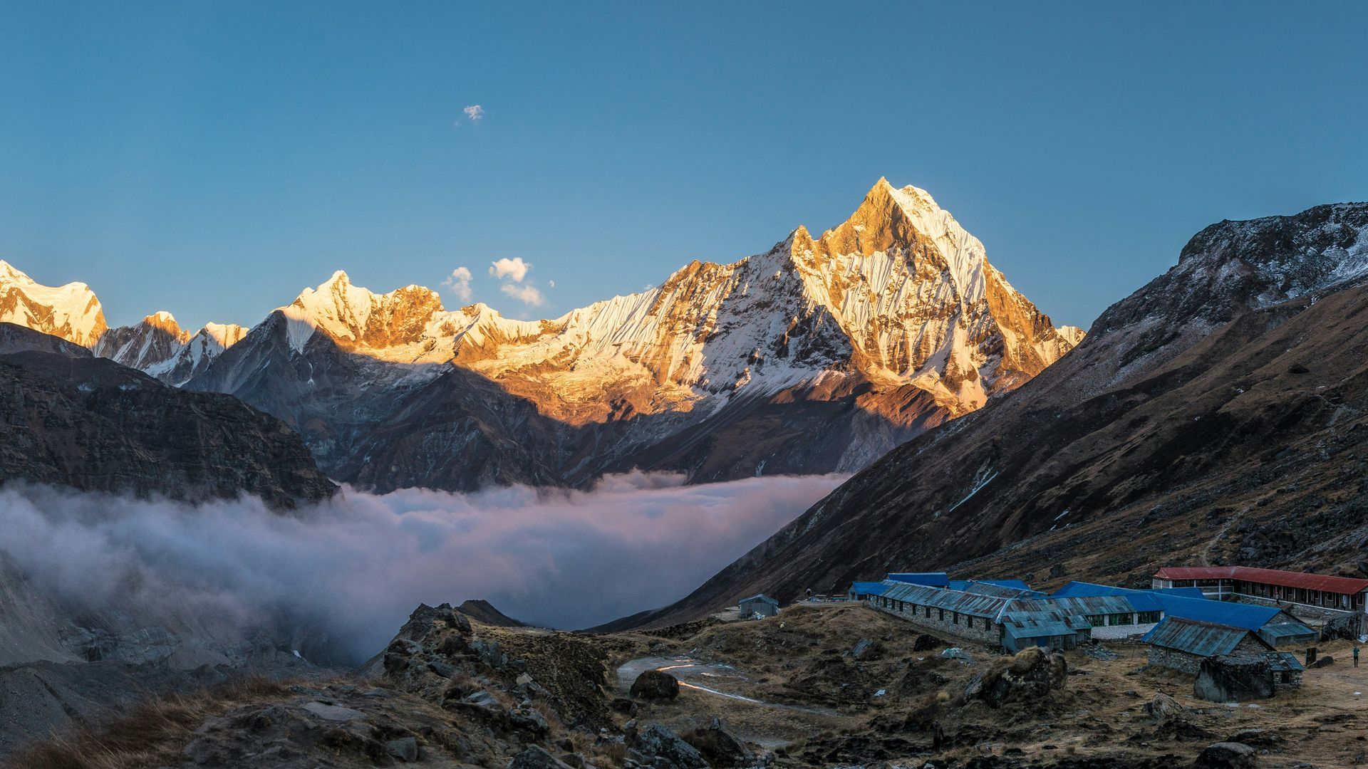 Snow-capped mountains at sunrise over a valley with clouds and a row of buildings in Annapurna, Nepal.