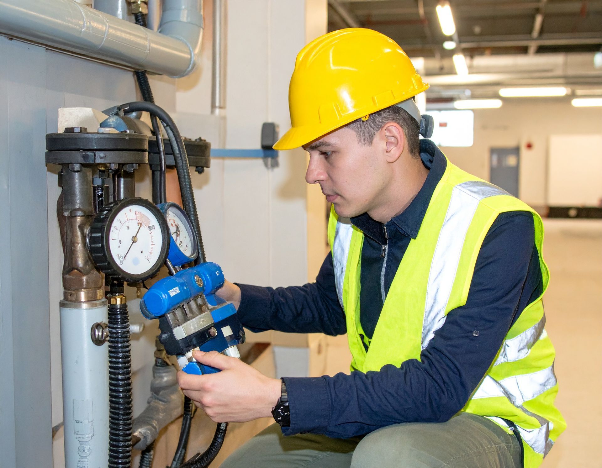 Technician in hard hat and safety vest inspecting industrial piping with a pressure gauge.