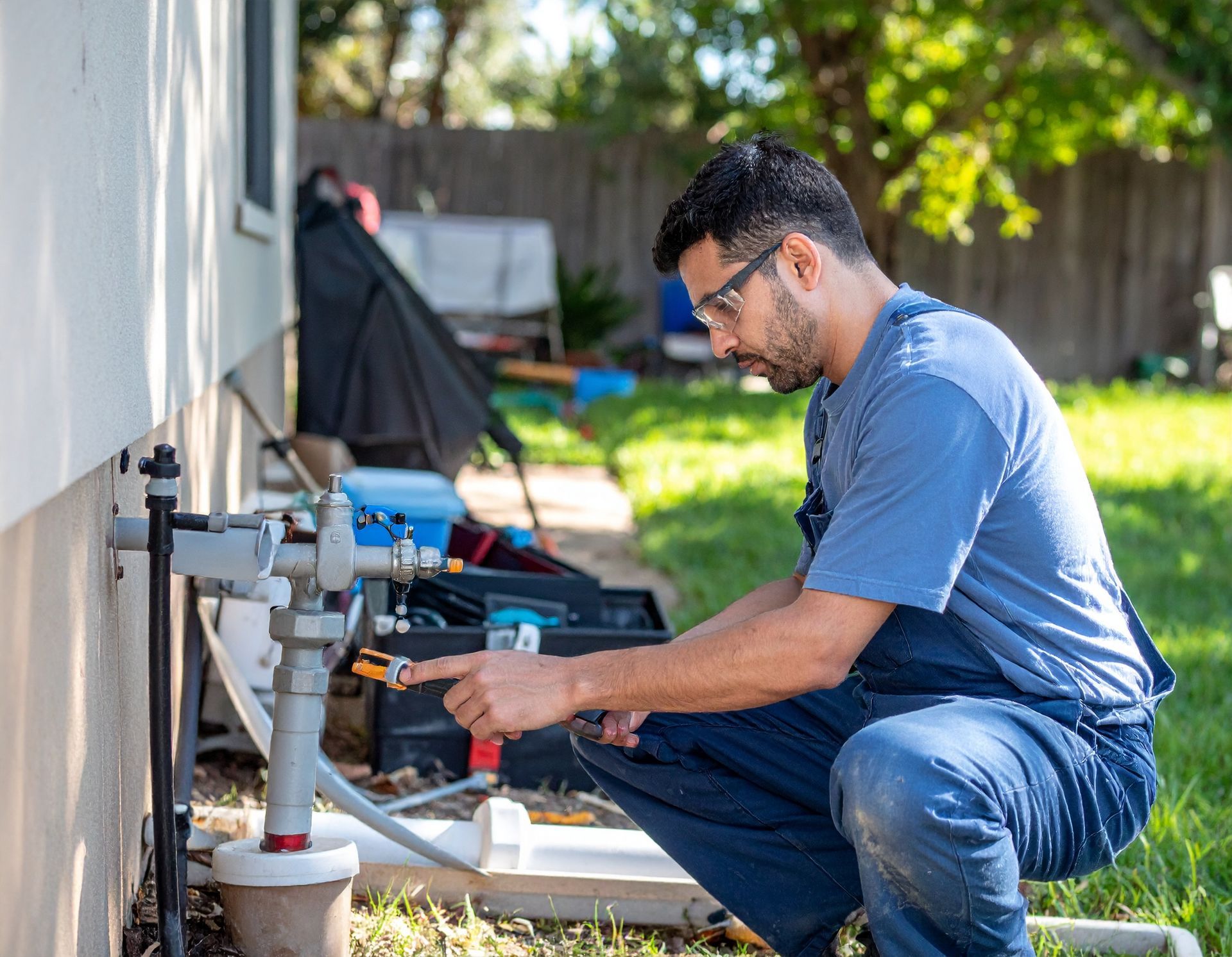 Technician in blue crouches beside outdoor HVAC unit, adjusting piping with tools in a sunny yard.
