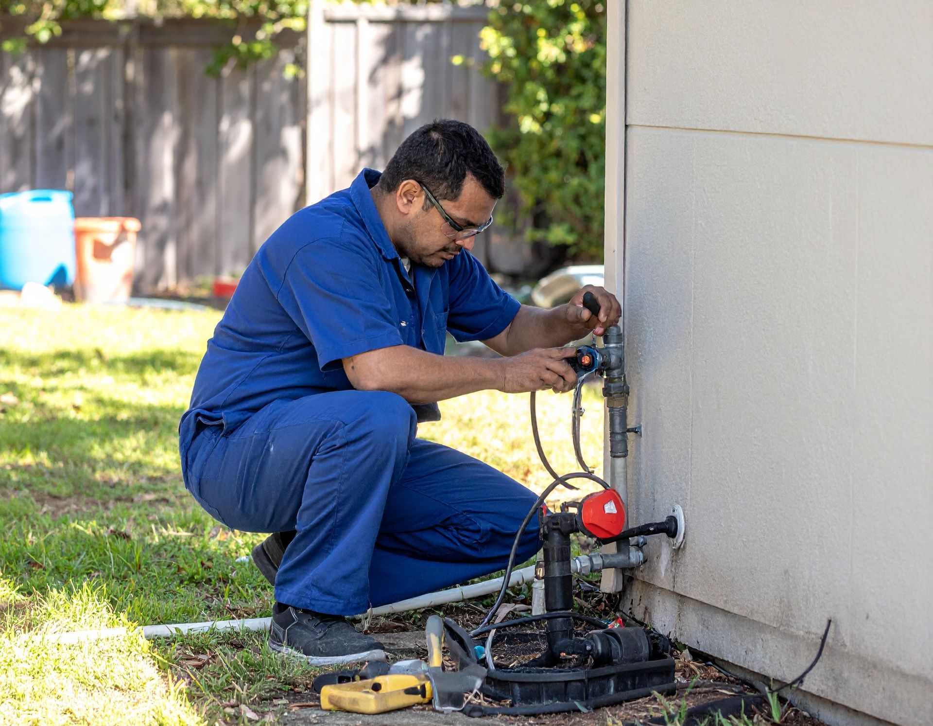 Technician in blue coveralls using plumbing equipment beside an outdoor wall.