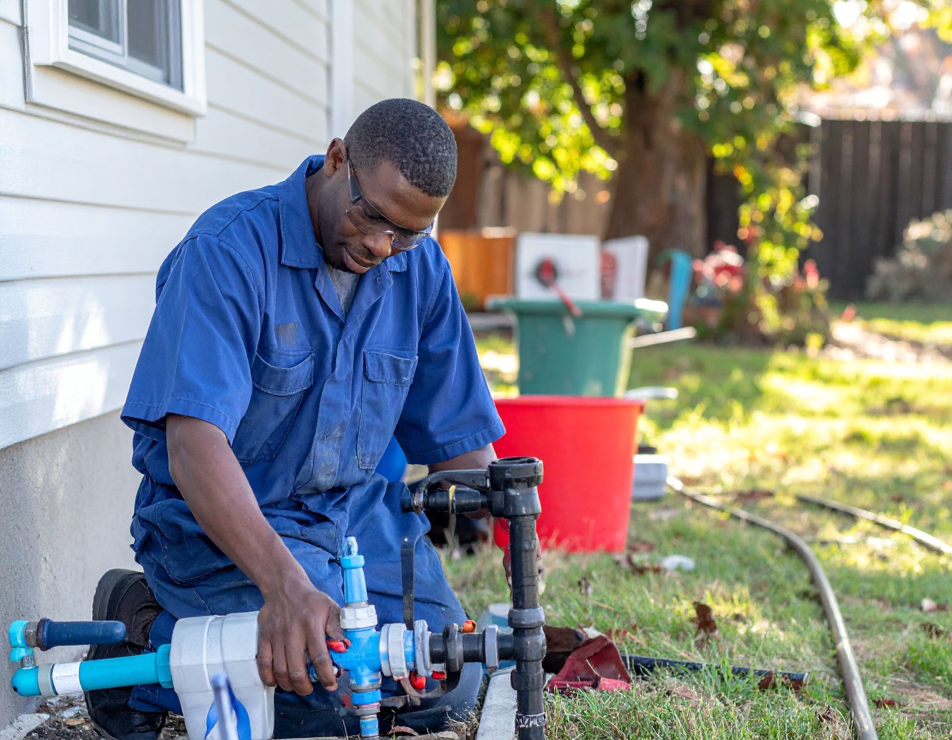 Person in blue workwear fixing outdoor plumbing by a house, with tools and buckets nearby