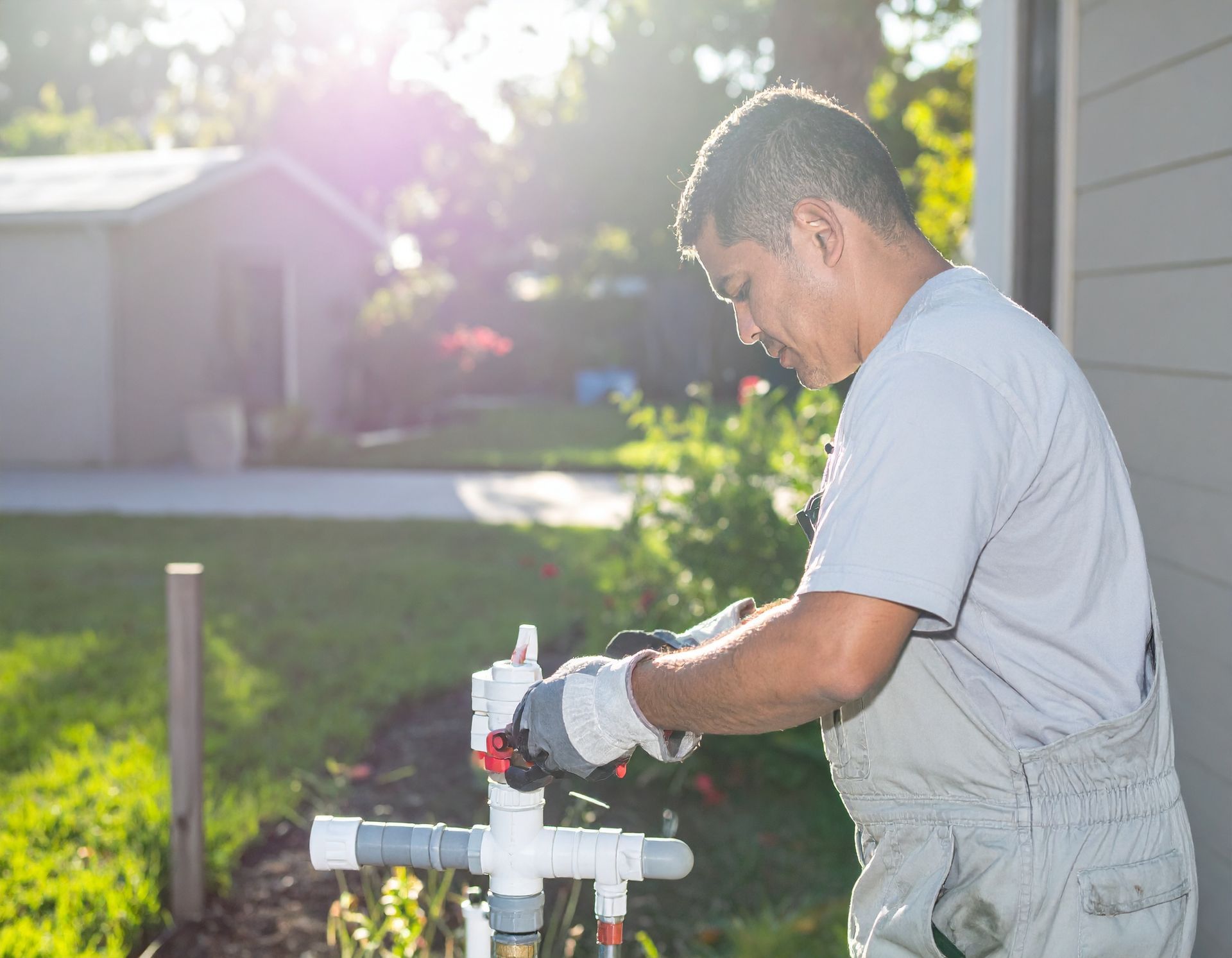Worker repairing outdoor water pipes in a sunny yard, wearing overalls and gloves