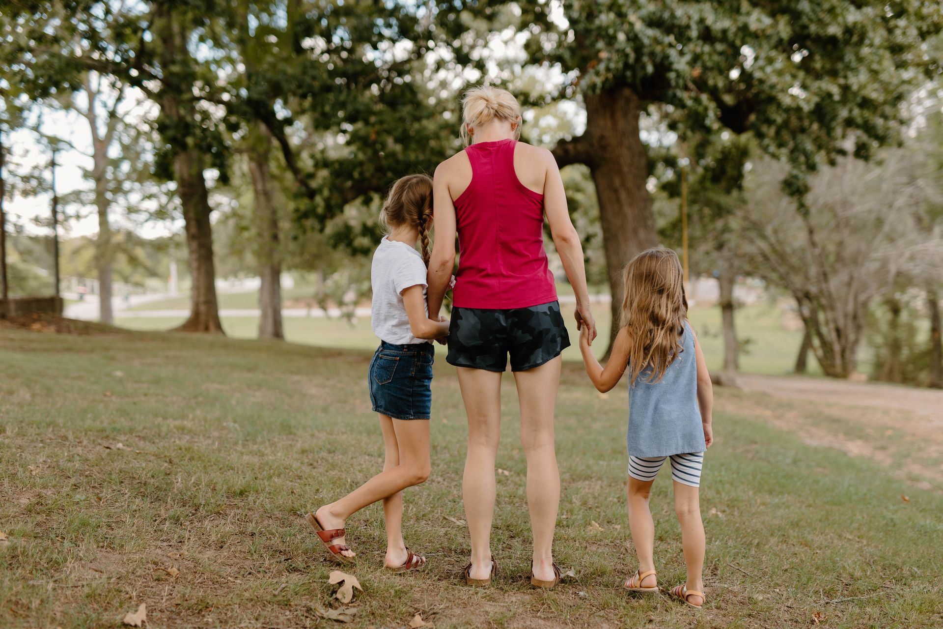 A woman and two little girls are walking in a park holding hands.