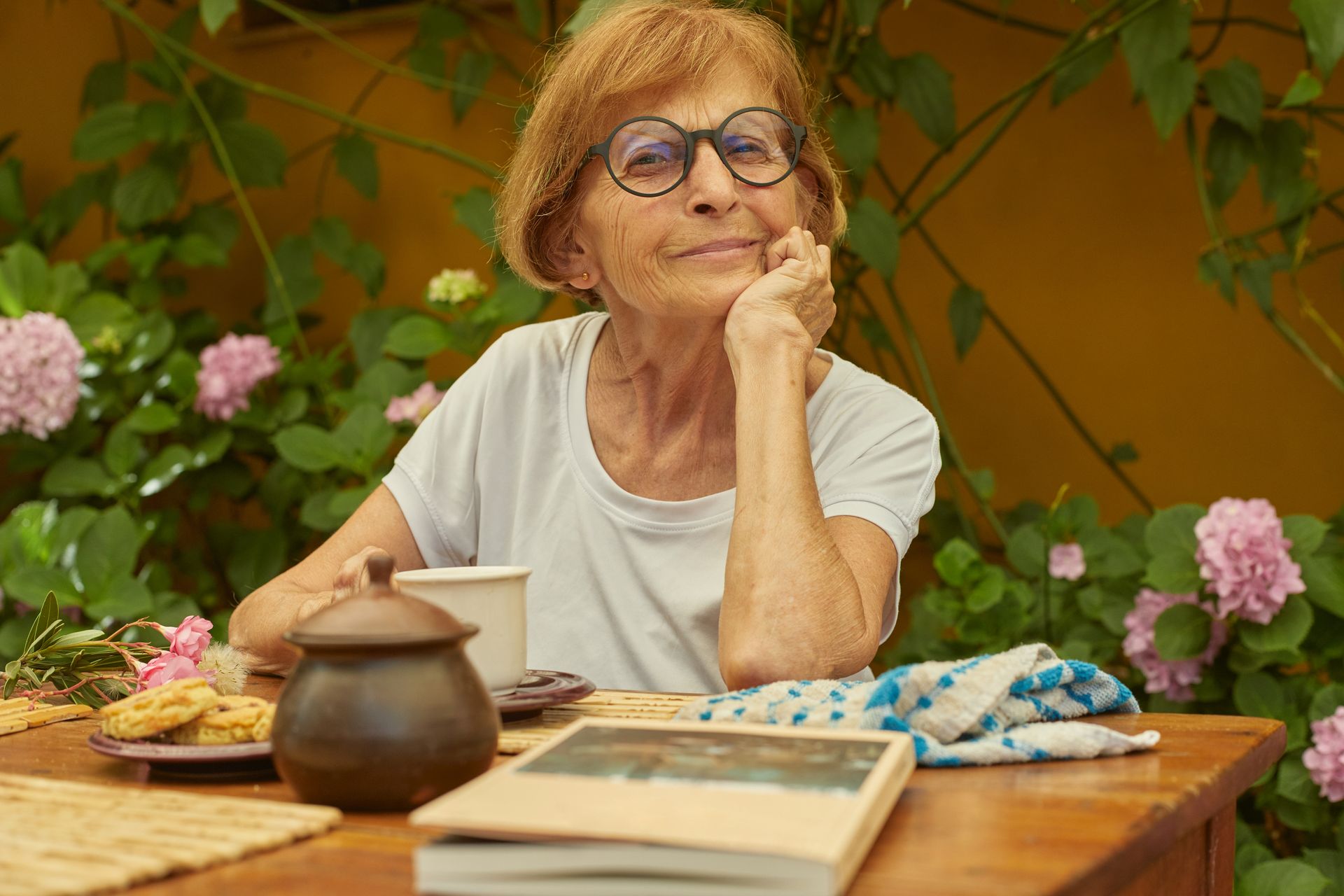An elderly woman is sitting at a table with a book and a cup of coffee.