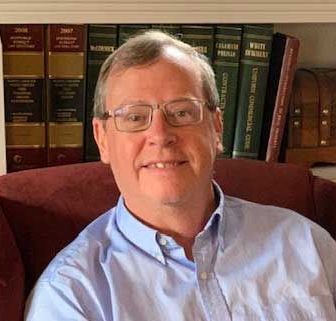 A man wearing glasses is sitting in a chair in front of a bookshelf.