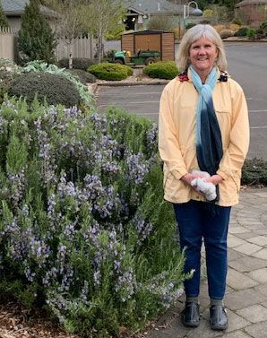 A woman in a yellow jacket is standing in front of a bush with purple flowers.