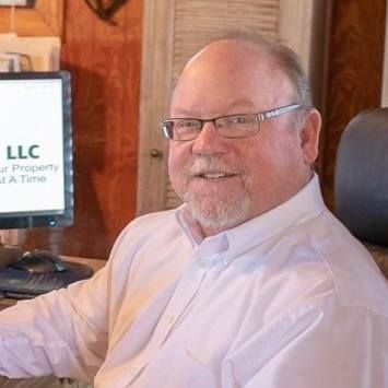 A smiling person with glasses wearing a white button-down shirt, sitting in an office chair in front of a computer.